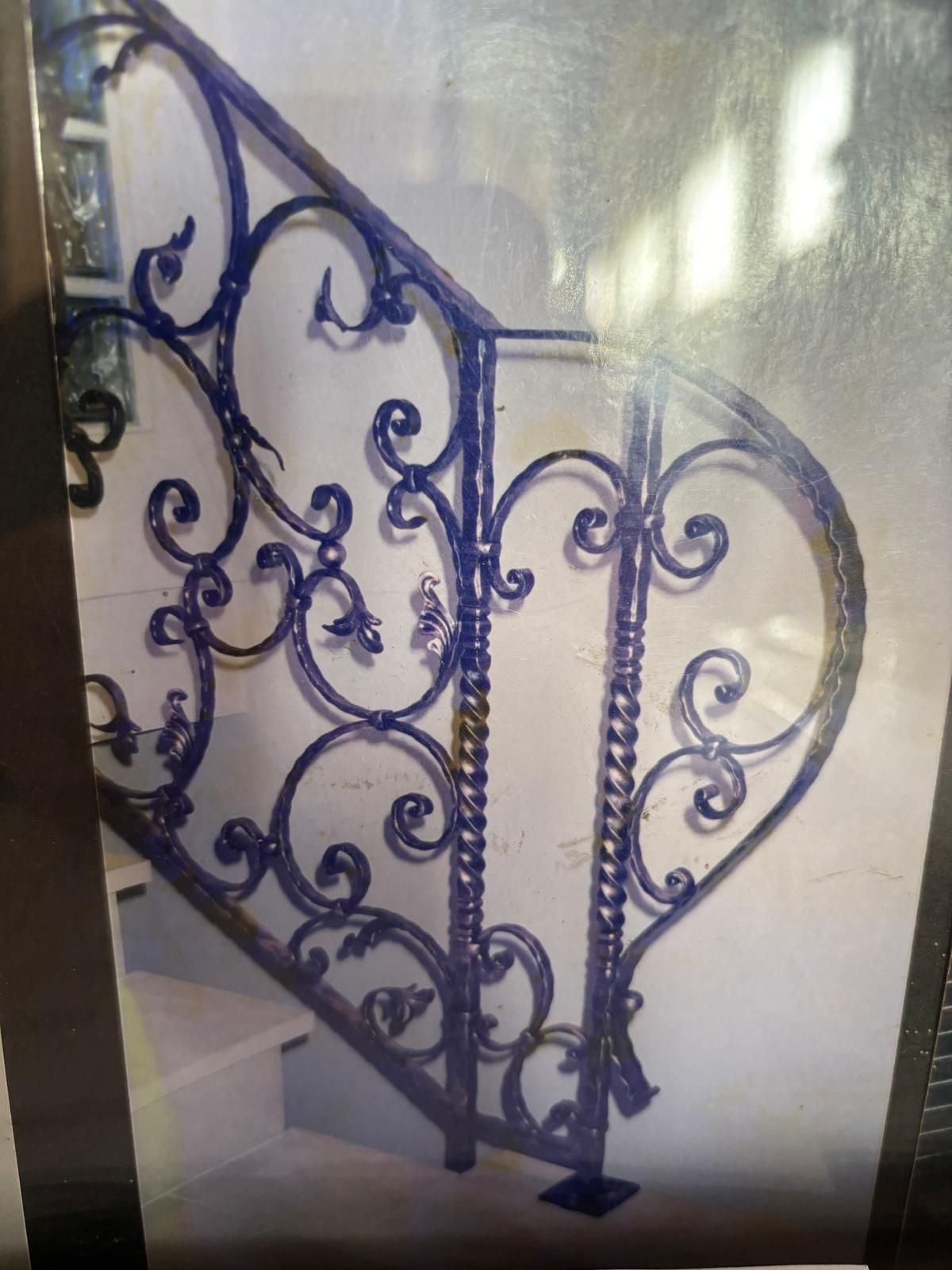 Ringhiera per scale in ferro battuto scuro con elaborate volute decorative e barre verticali ritorte su una parete di colore chiaro.