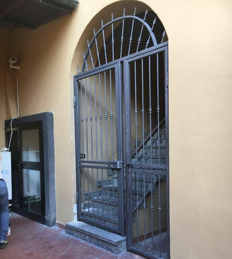 Un cancello di sicurezza ad arco in metallo scuro, installato in una parete color beige di un edificio, con una scala in pietra visibile all'interno dell'ingresso.