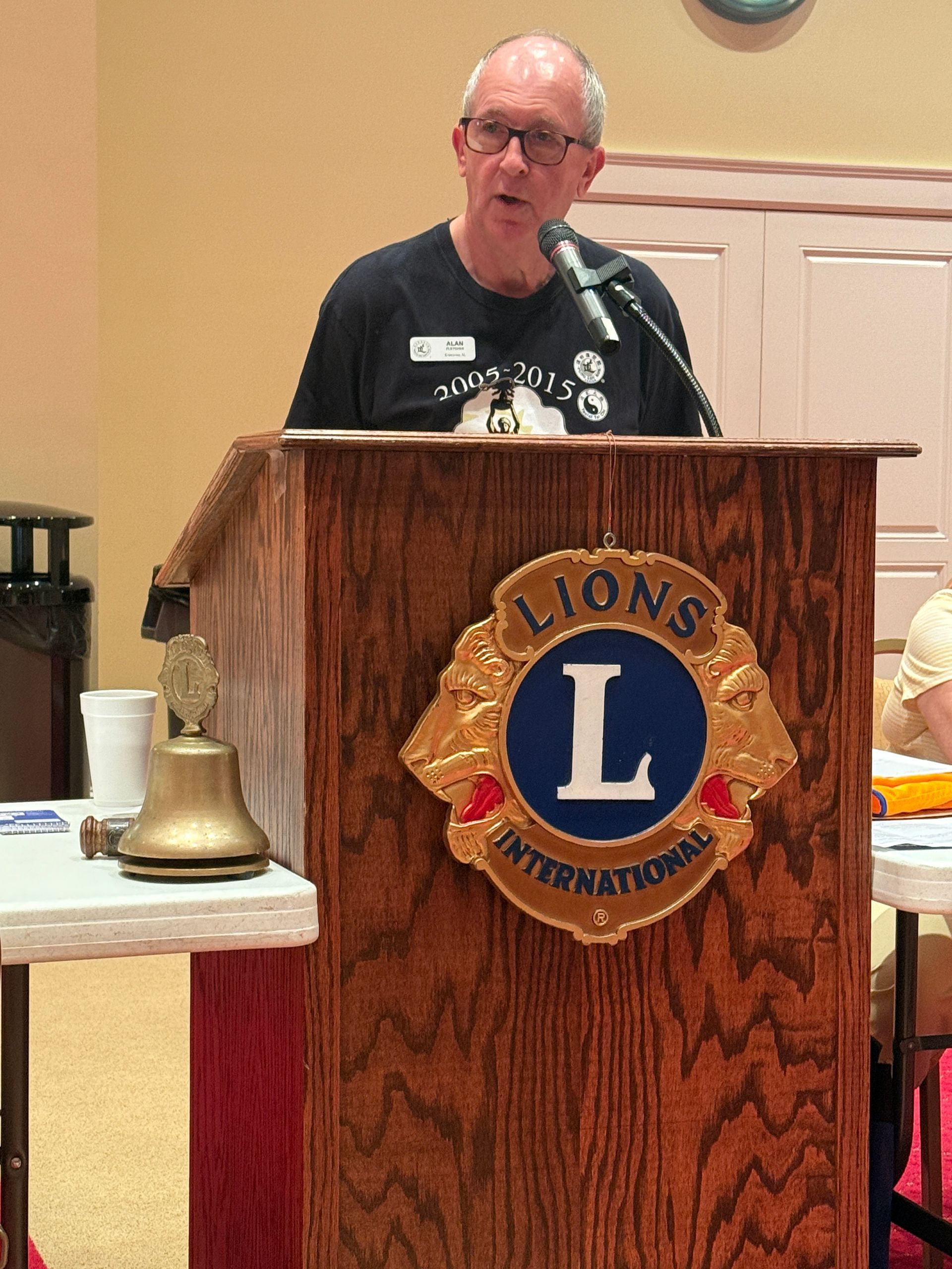 Man speaking at a Lions Club podium, wearing a black shirt, inside a meeting room.