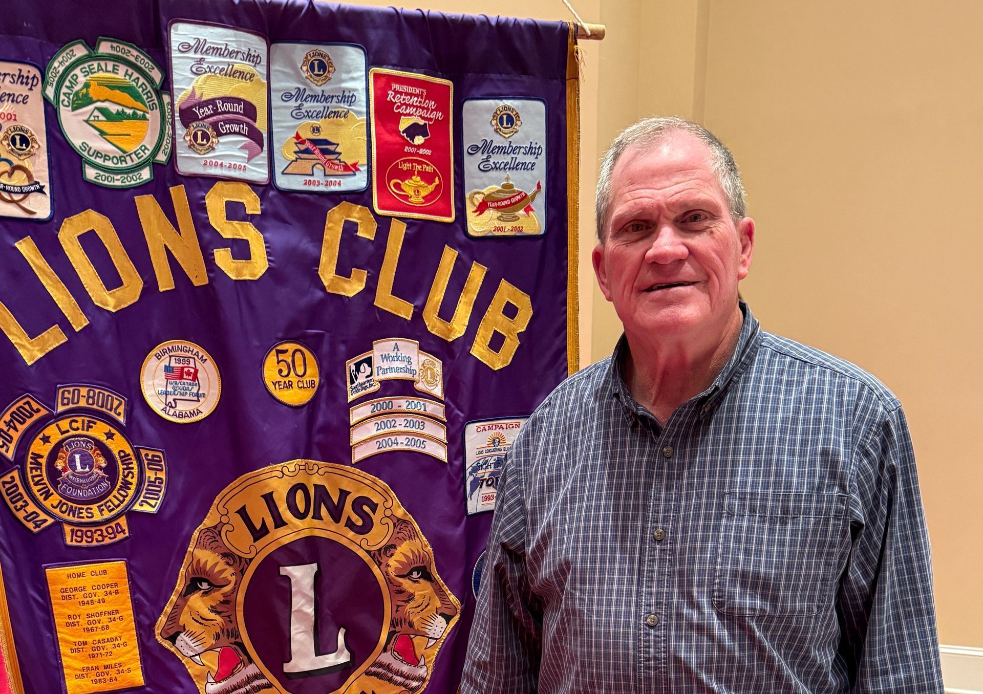 Man in blue shirt stands next to a Lions Club banner with patches.