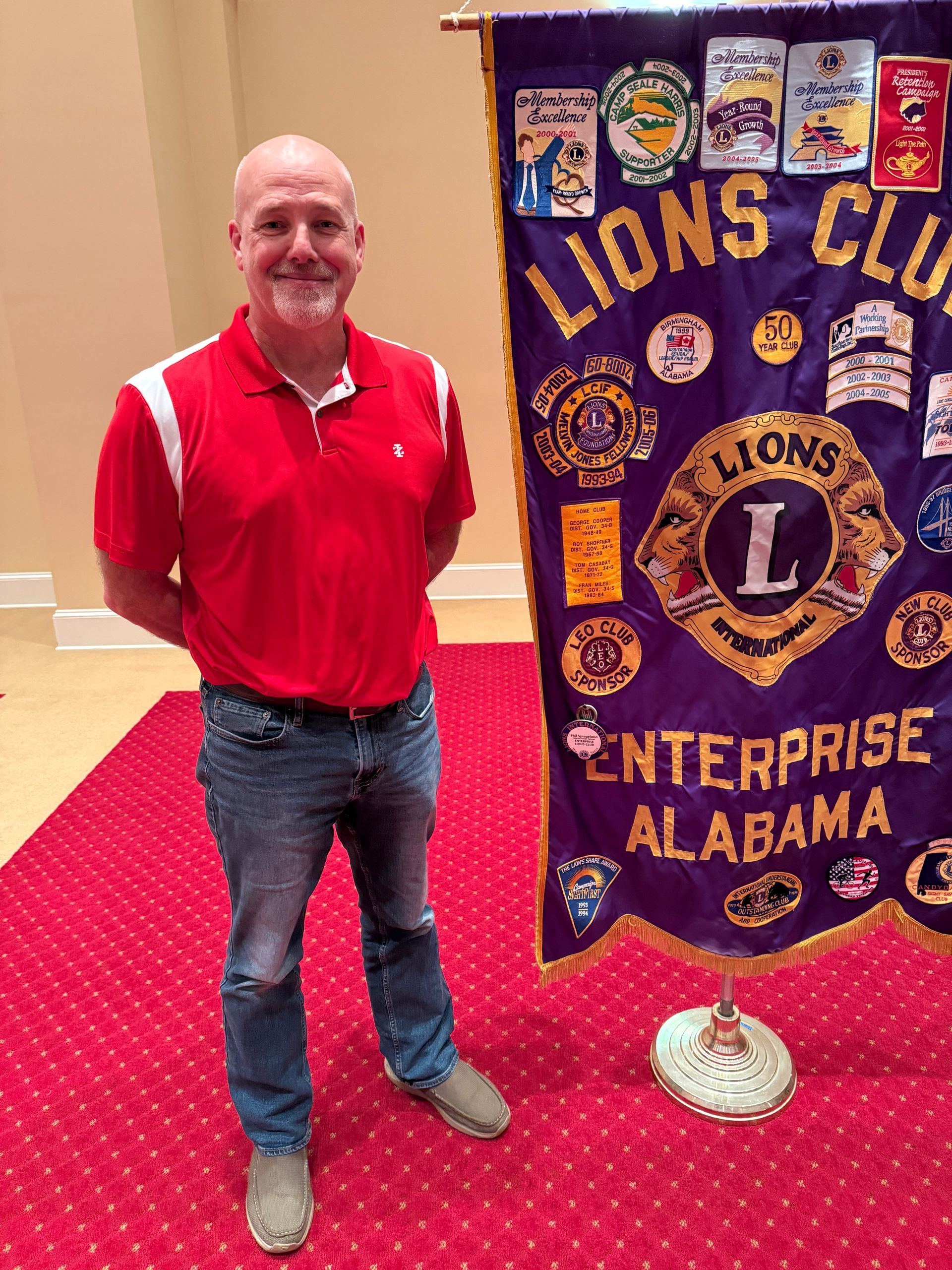 Man in red shirt and jeans stands next to a purple Lions Club banner in Enterprise, Alabama.