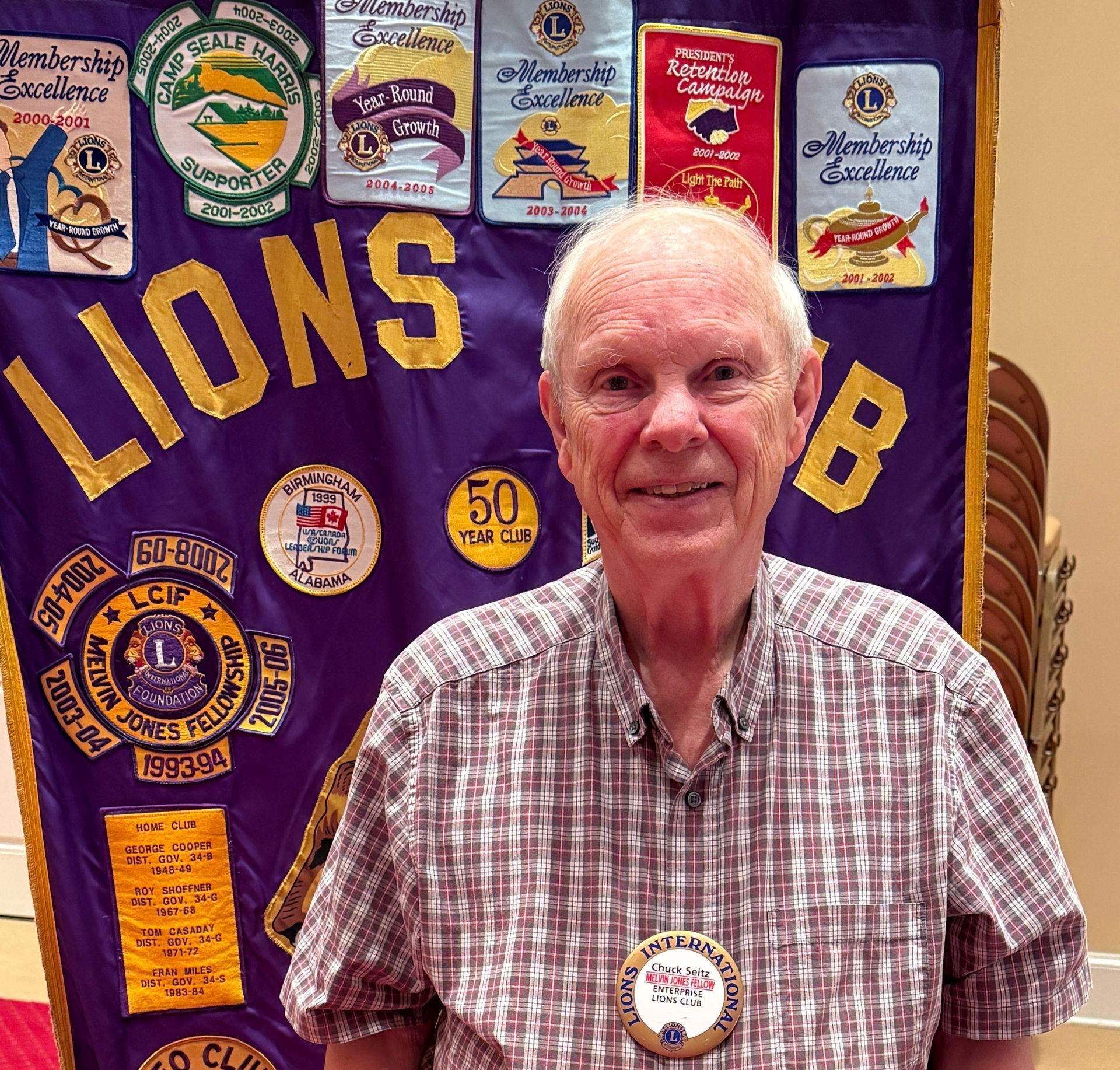 Older man in checkered shirt stands before a purple Lions Club banner with patches.
