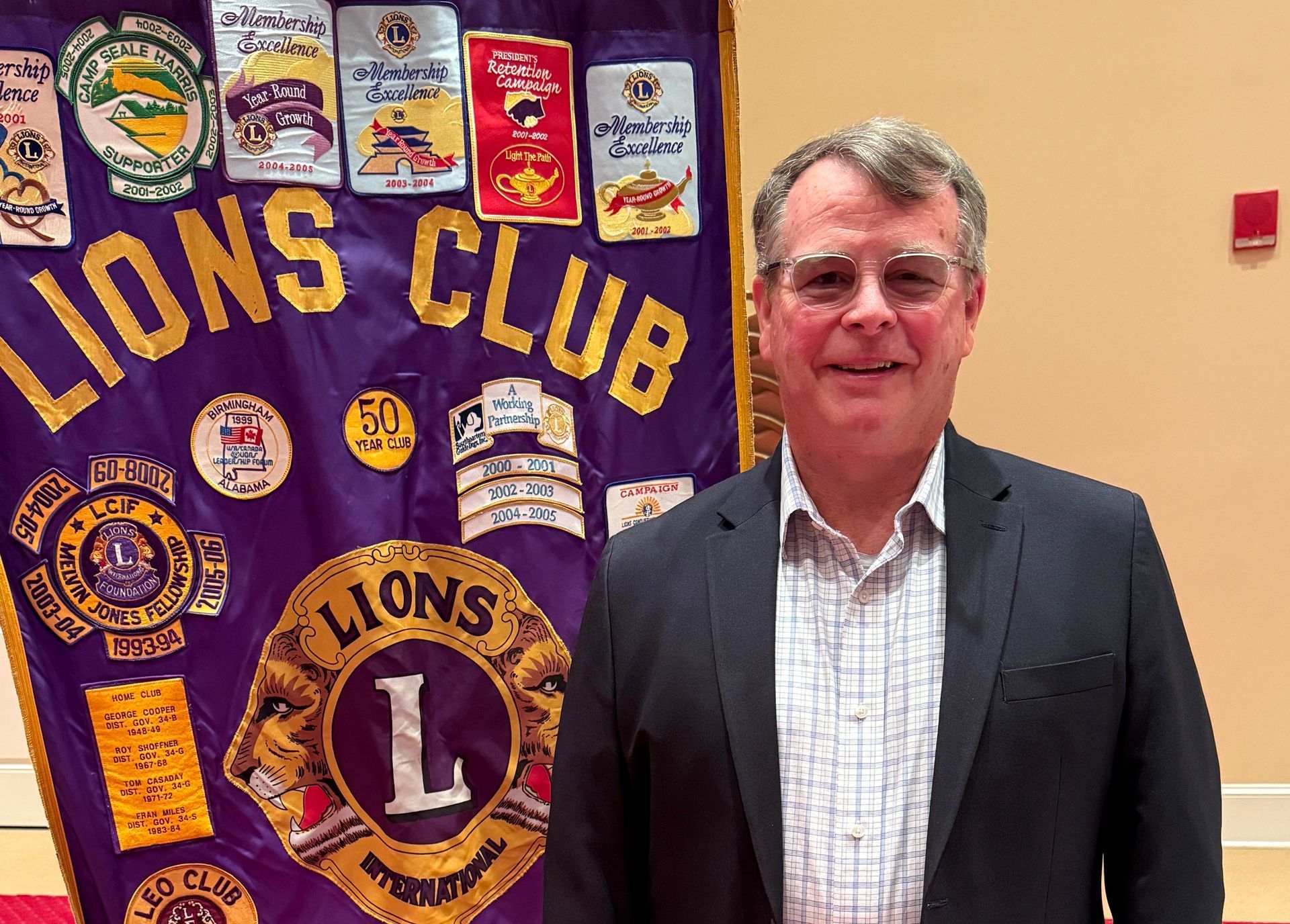 Man in suit stands in front of Lions Club banner; smiles.
