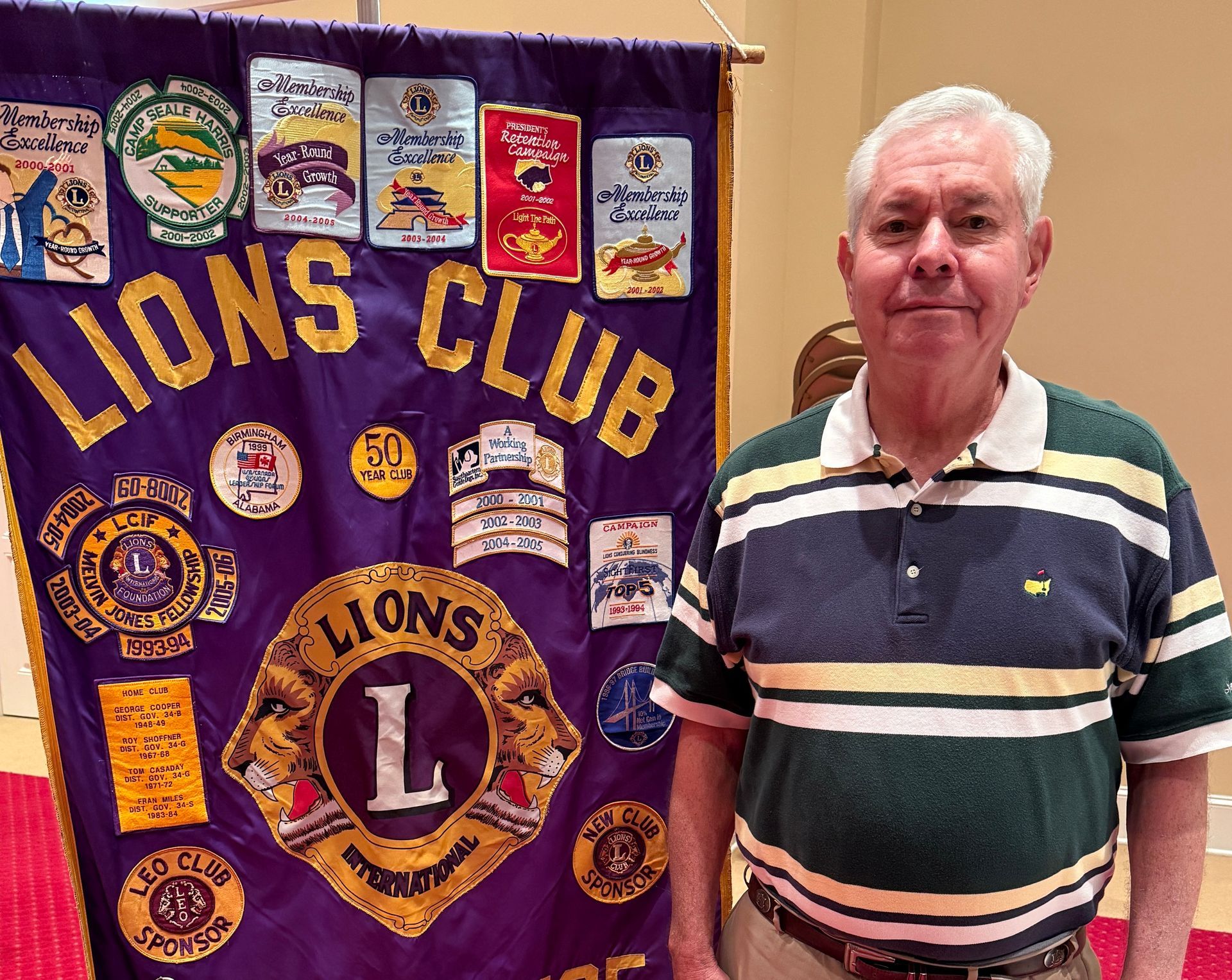 Man stands beside a Lions Club banner. He wears a green and white striped shirt and khaki pants.