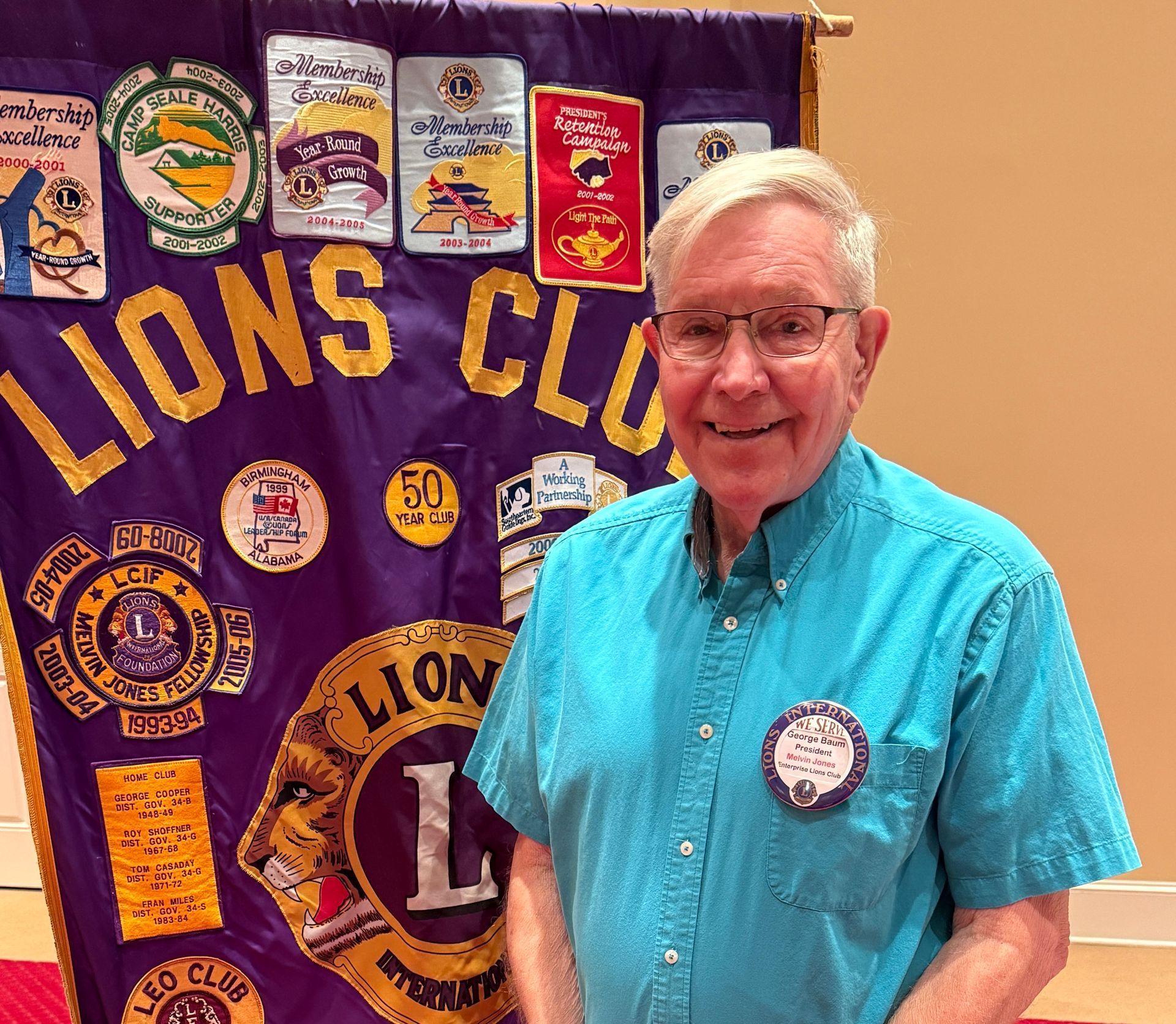 Man in blue shirt stands in front of a Lions Club banner. He wears glasses and a name badge.