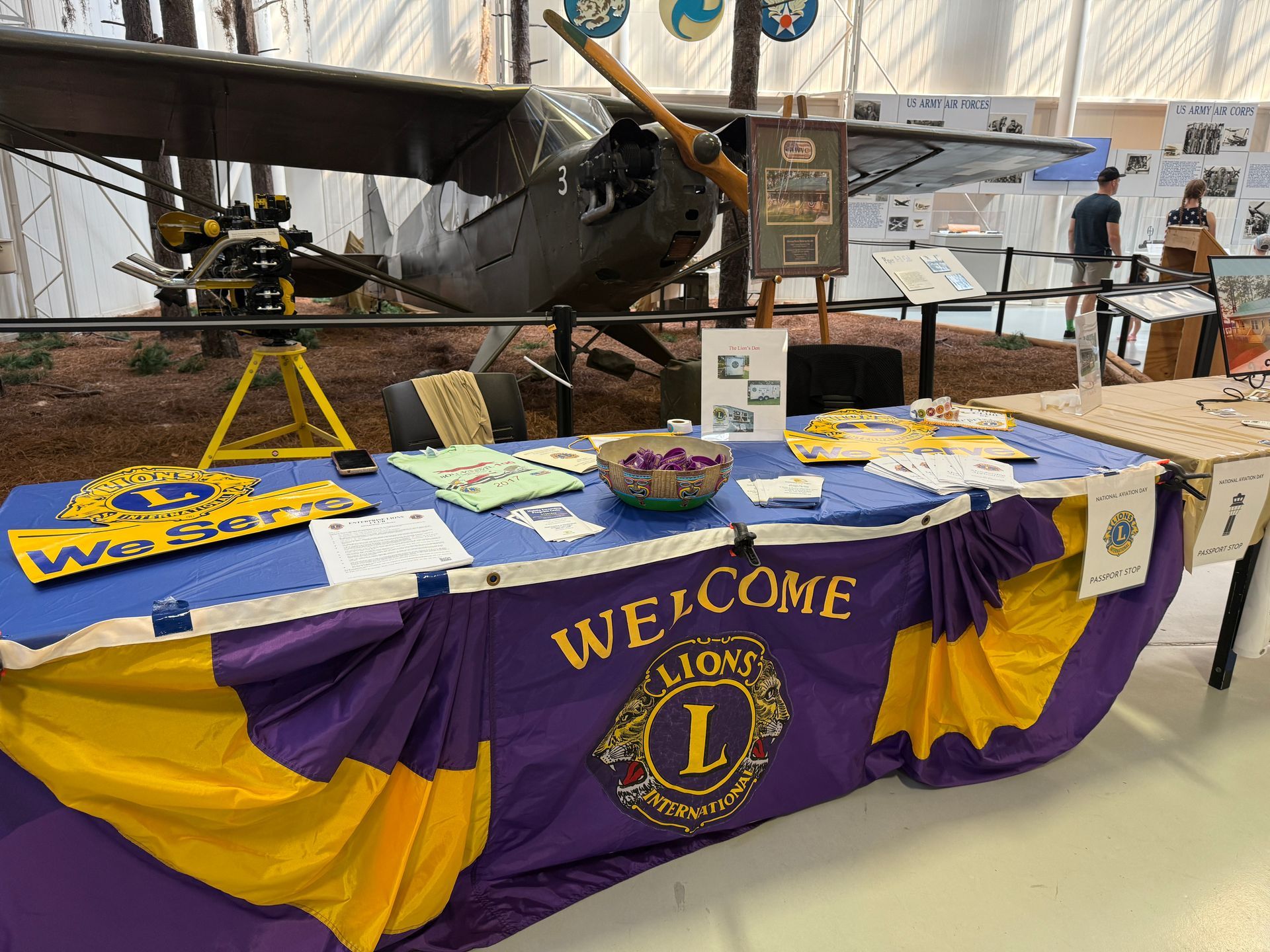 Lions Club welcome table at an aviation museum, purple and gold banner, plane in background.