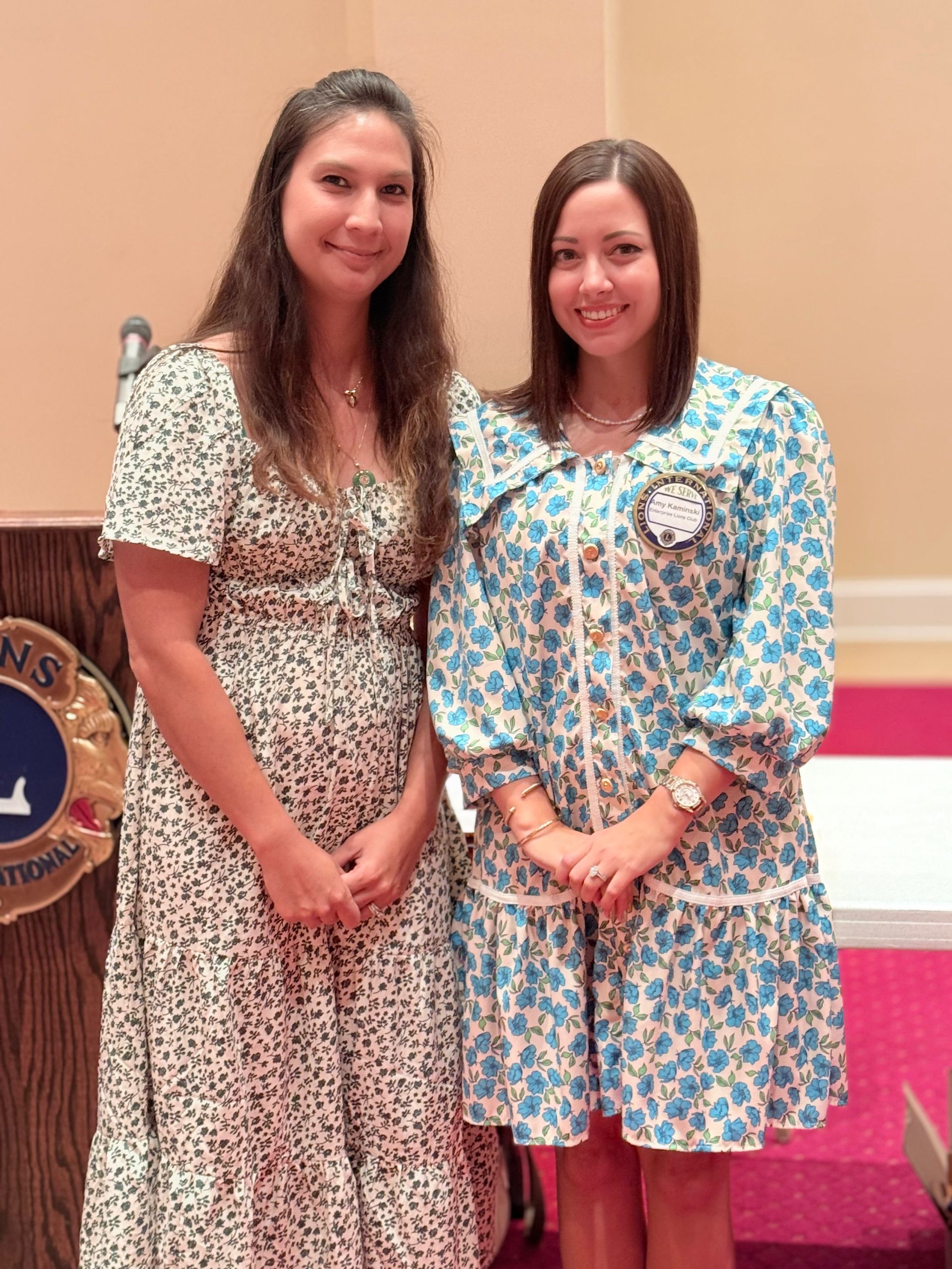 Two women stand by a podium. One wears a floral dress, and the other a blue floral dress.