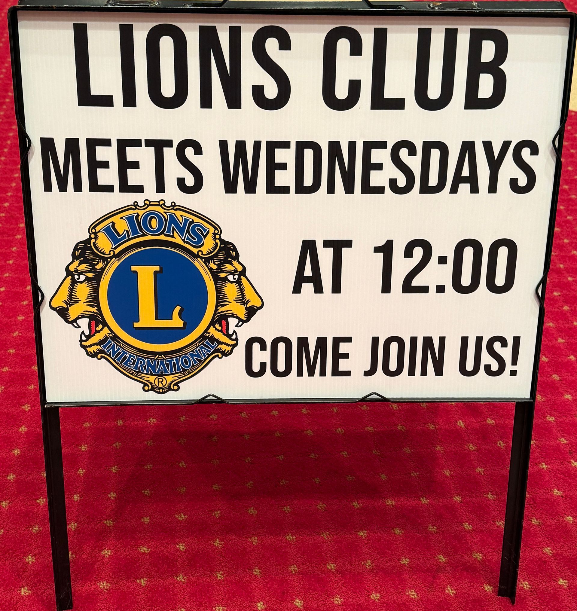 Sign: Lions Club meets Wednesdays at 12:00. Come join us! Features club logo, black text on white, red carpet.
