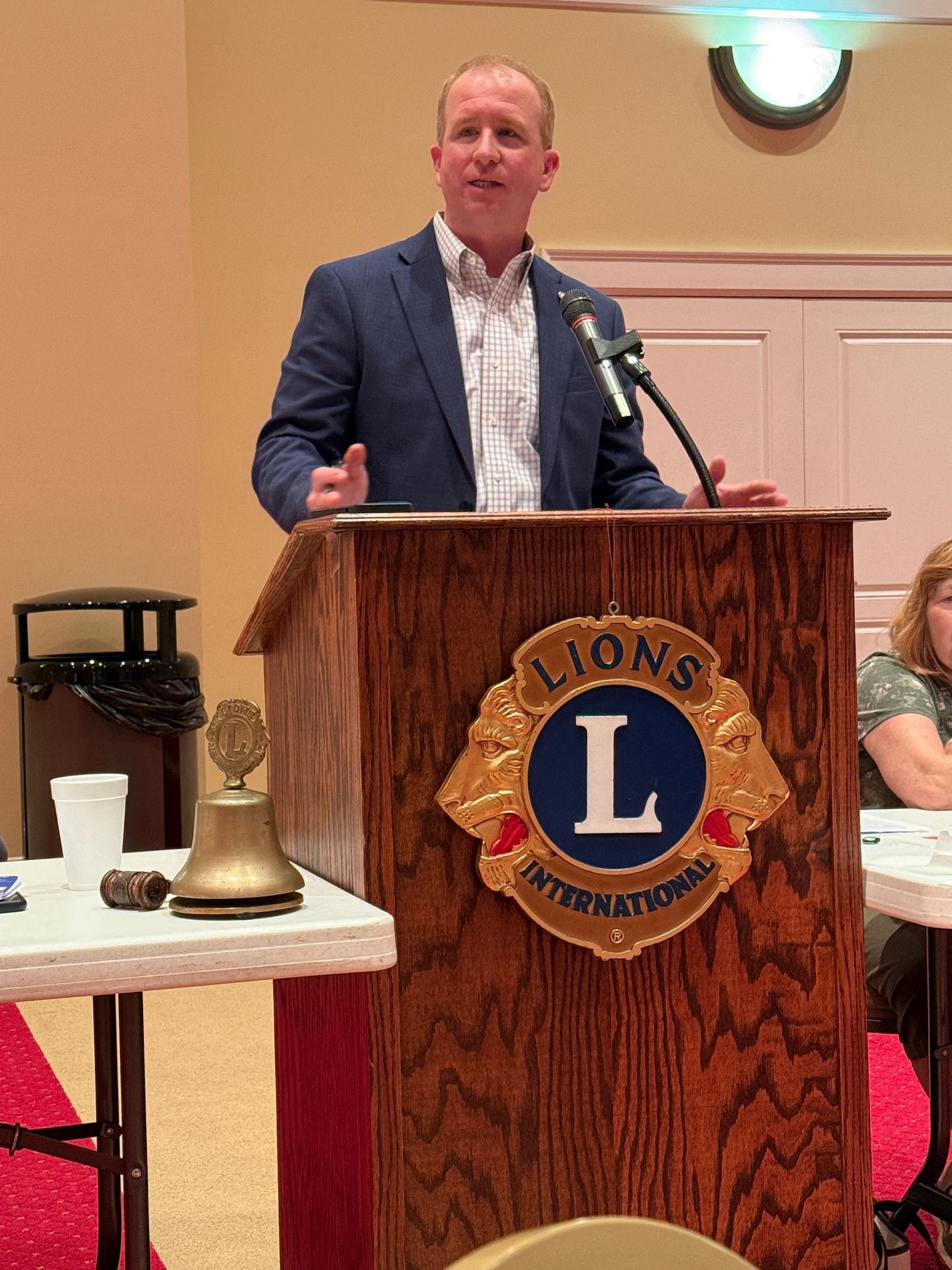 Man speaking at a Lions Club podium. He wears a blue jacket. The podium has a Lions Club emblem.