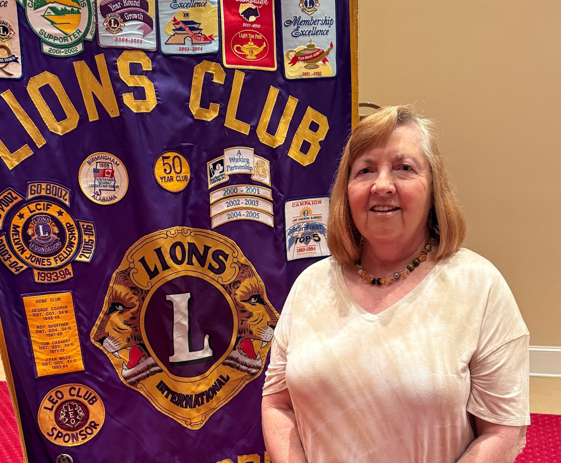 Woman standing in front of a Lions Club banner, smiling, with light-colored clothing.
