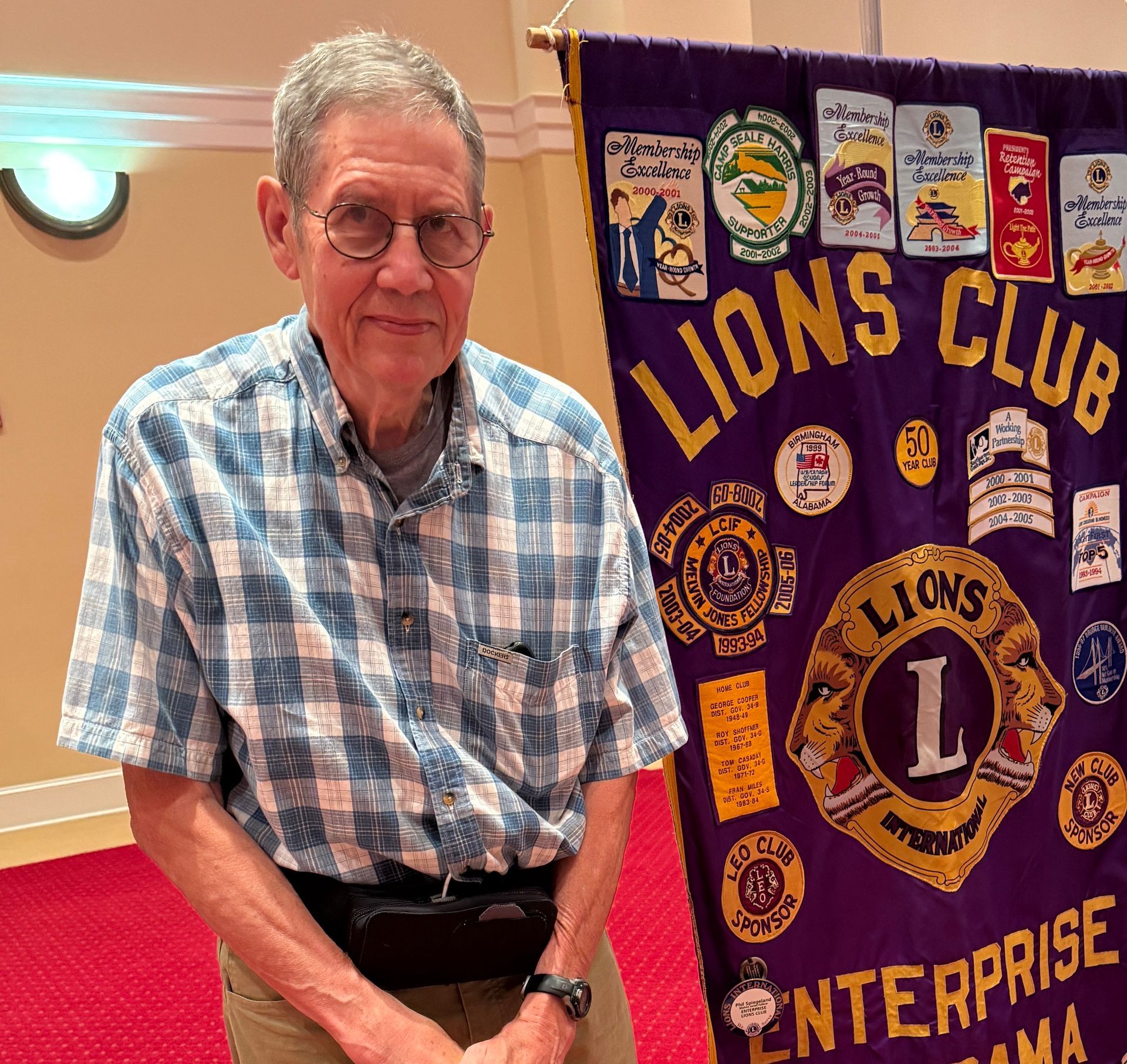Man in glasses and plaid shirt stands next to a purple Lions Club banner.