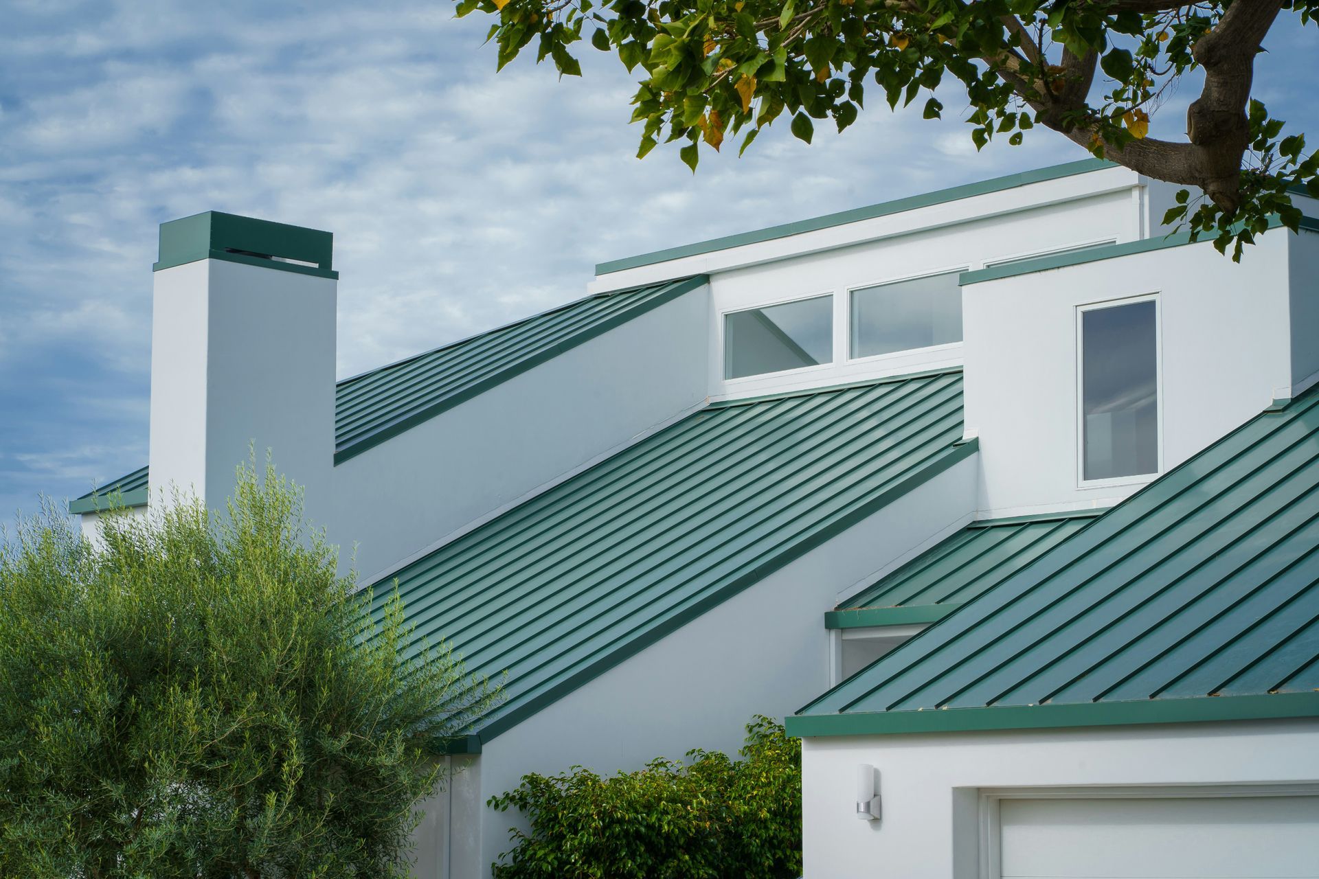 White house with green metal roofs, chimney, and trees under a blue sky