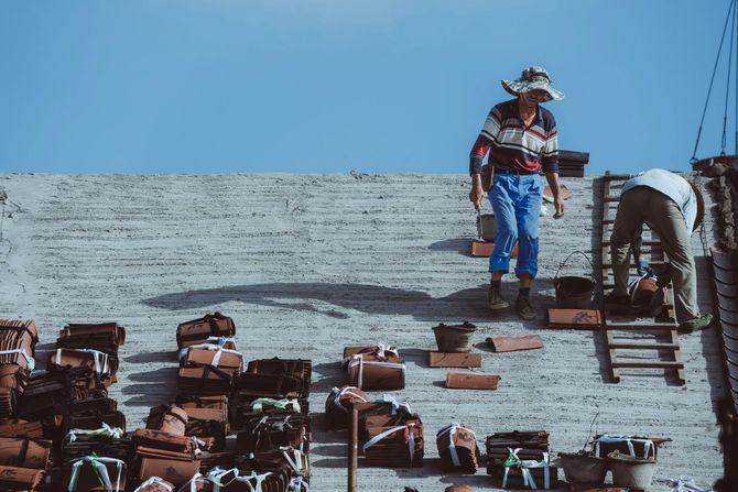 Workers on a rooftop installing red tiles, with one person standing and another bending over under a clear blue sky