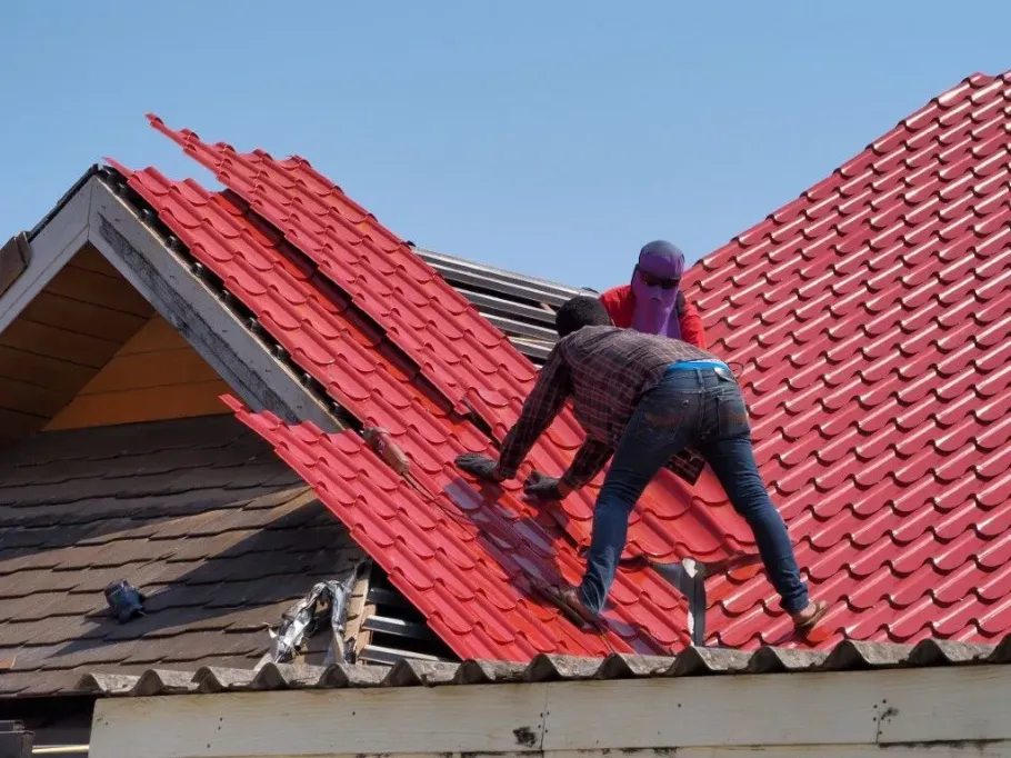 Two workers installing red roof tiles on a house roof under a blue sky