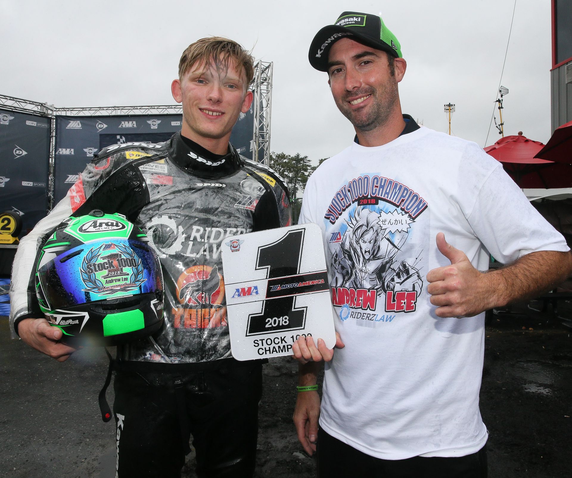 A young man in racing gear holds a trophy with another man. Both are smiling and outside.