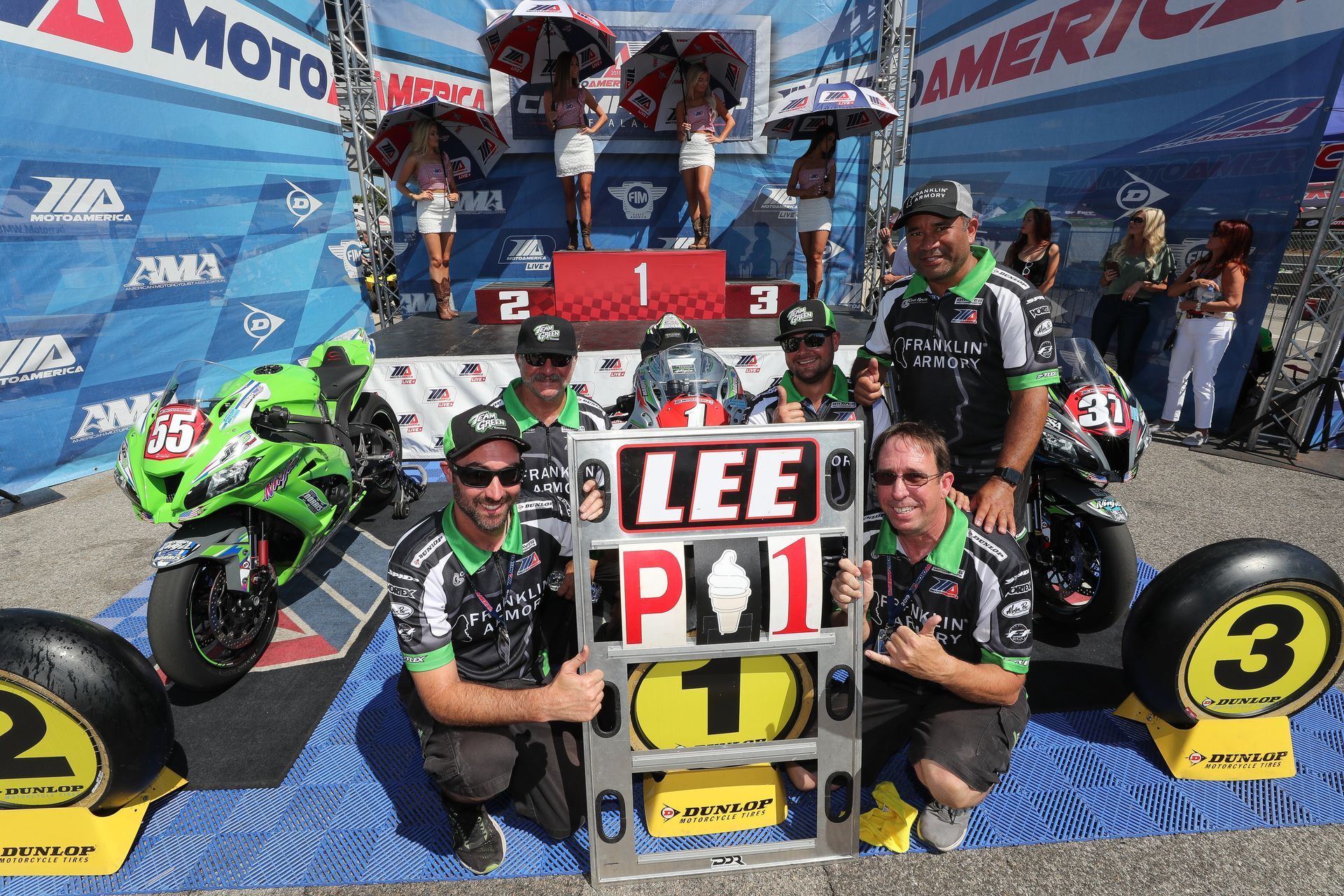 Motorcyclist Lee on podium, Kawasaki bike, crew, victory celebration, trophies, track, sunny.