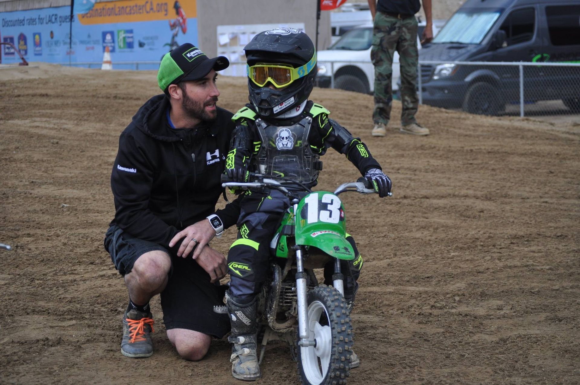 A child in motocross gear sits on a small motorcycle with an instructor in an outdoor setting.