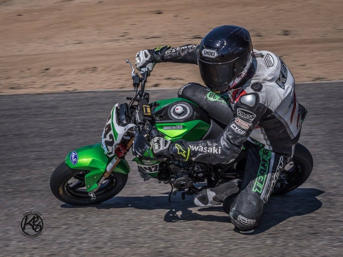 Motorcycle racer leans low on a green and black bike on a track, wearing a black helmet and racing suit.