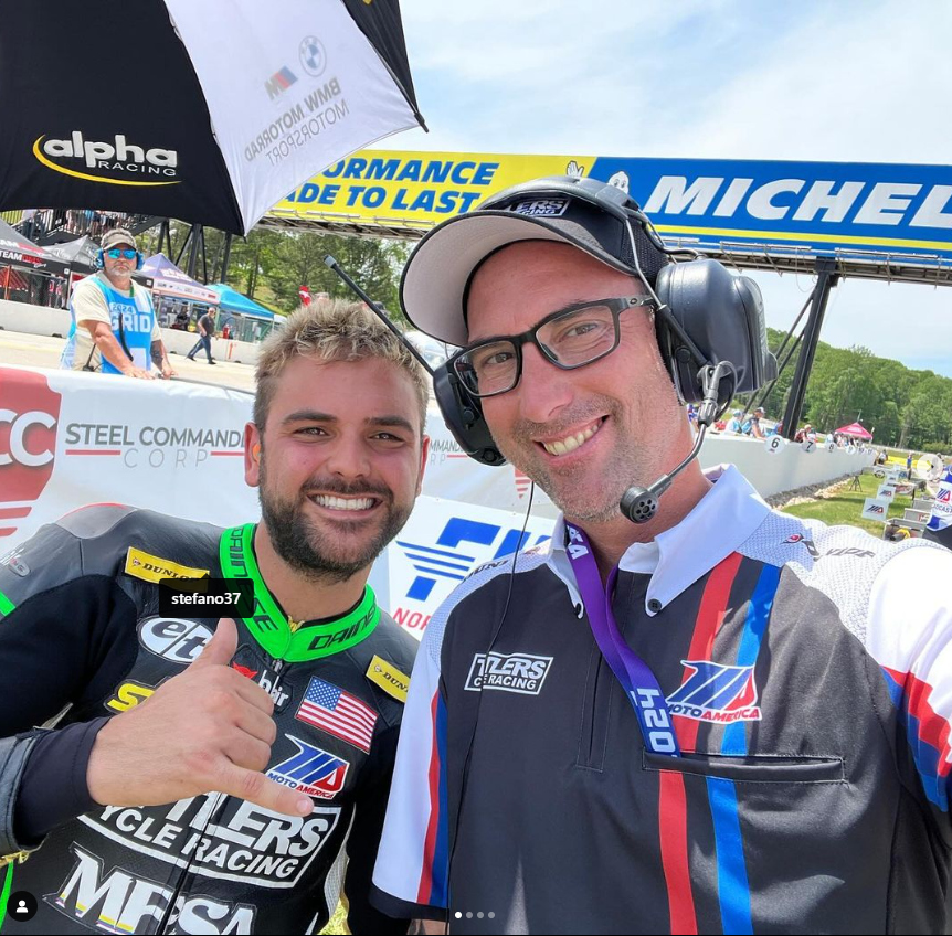 Two men smiling at a racetrack. One wears a racing suit, gives a thumbs up; the other, a headset and team shirt.