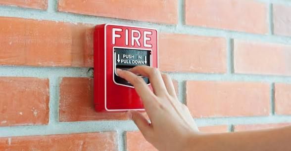 Red fire alarm on a tan concrete pillar, with clear plastic cover and metal conduit.