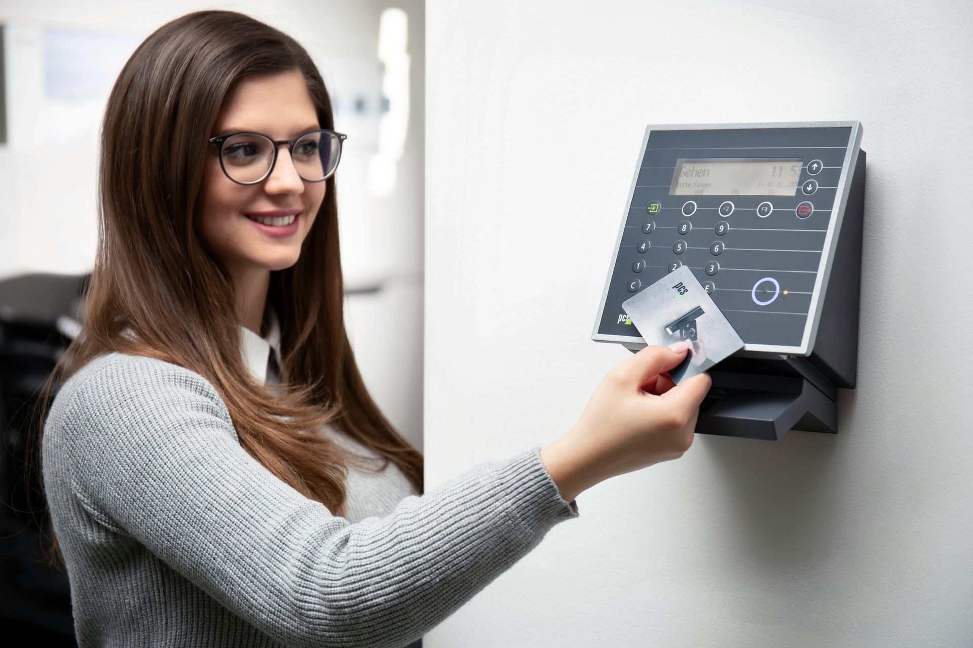 Woman swiping ID card at a time clock on a white wall. She wears glasses and smiles.