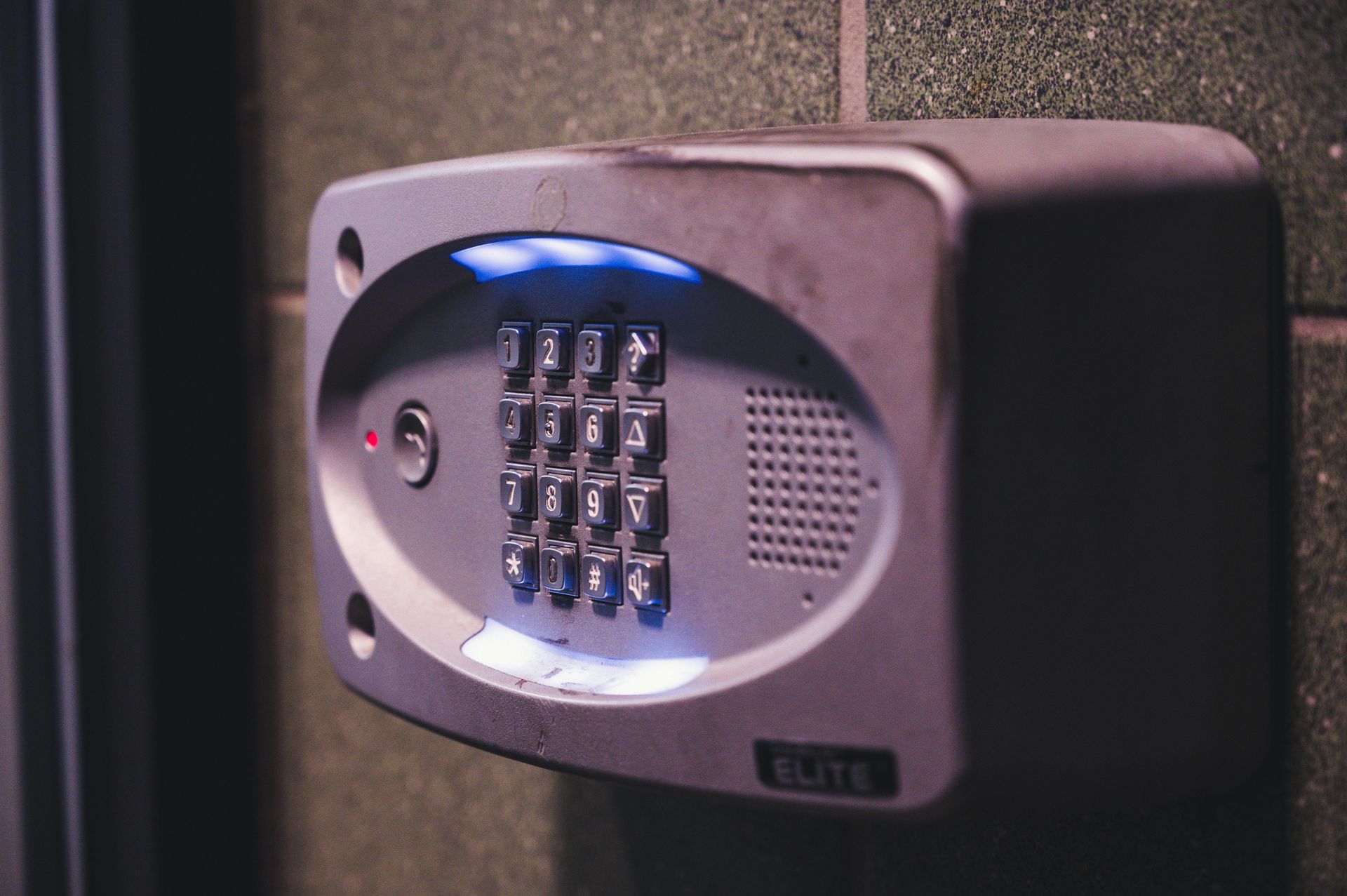 Gray intercom keypad with illuminated buttons mounted on a dark green wall.