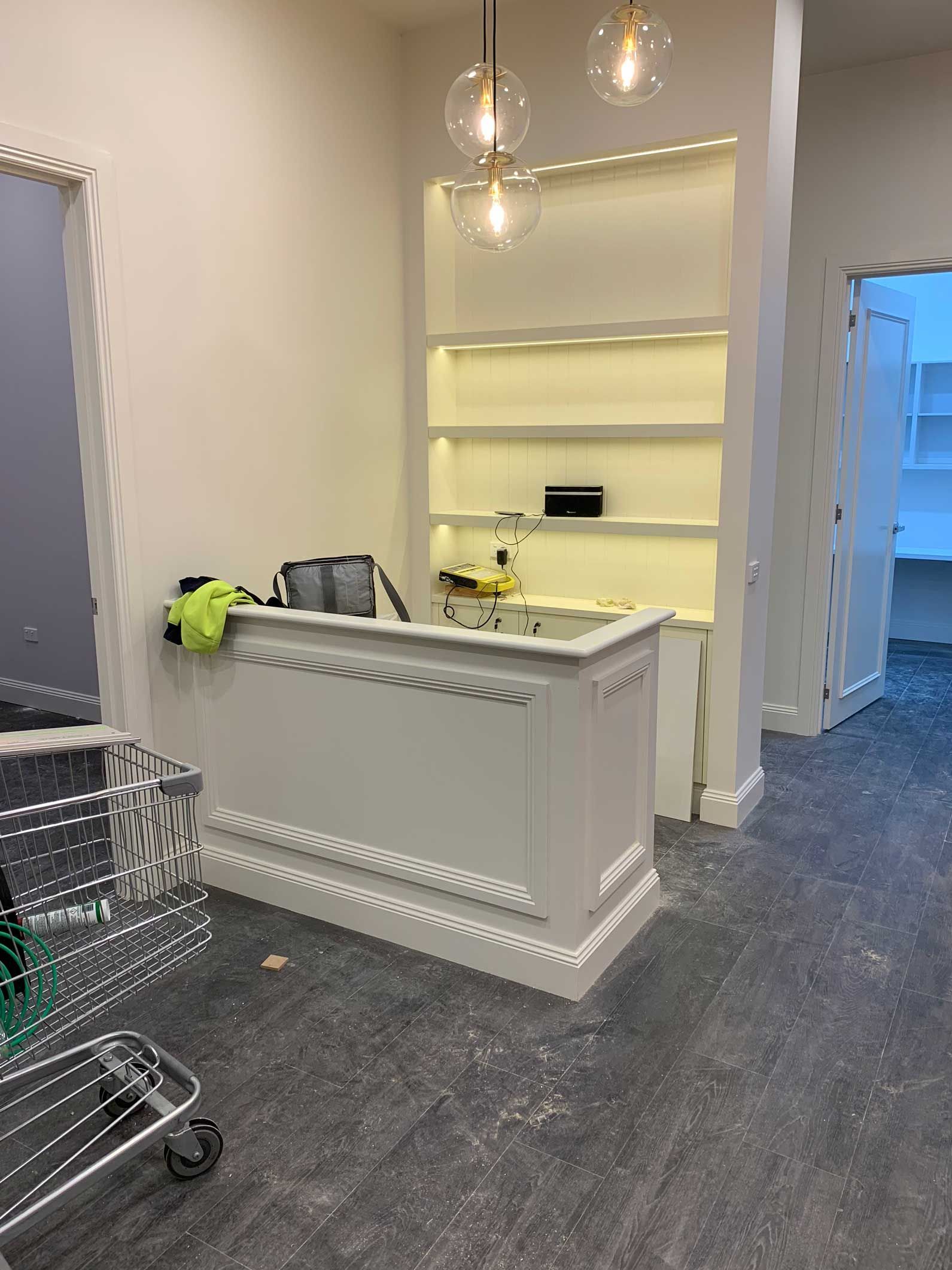 White reception desk with decorative trim and recessed shelves, lit by overhead globe lights, in a cream-colored room.