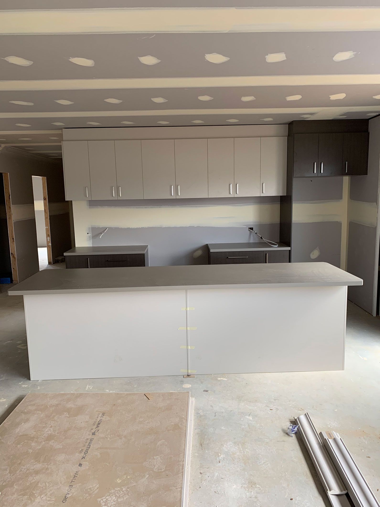 Kitchen under construction with cabinets and island. Drywall walls, unfinished floors, and a countertop.