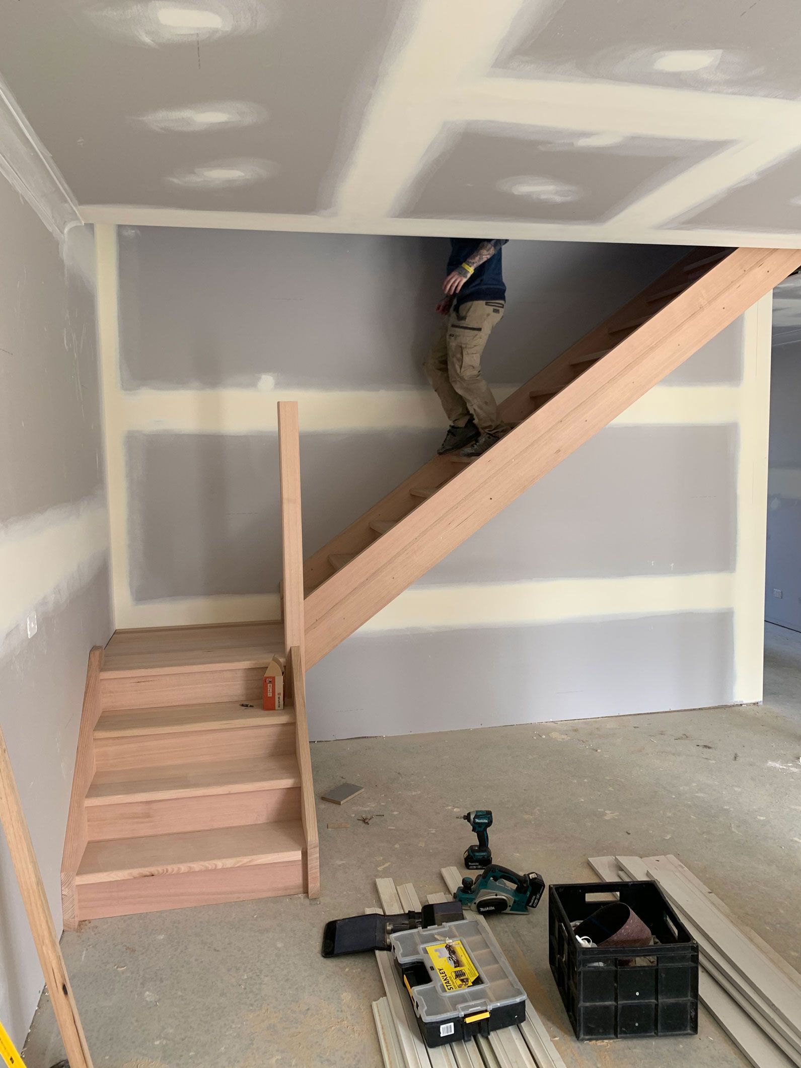 A person walks up a wooden staircase in a room under construction. Drywall lines the walls, tools are visible on the floor.
