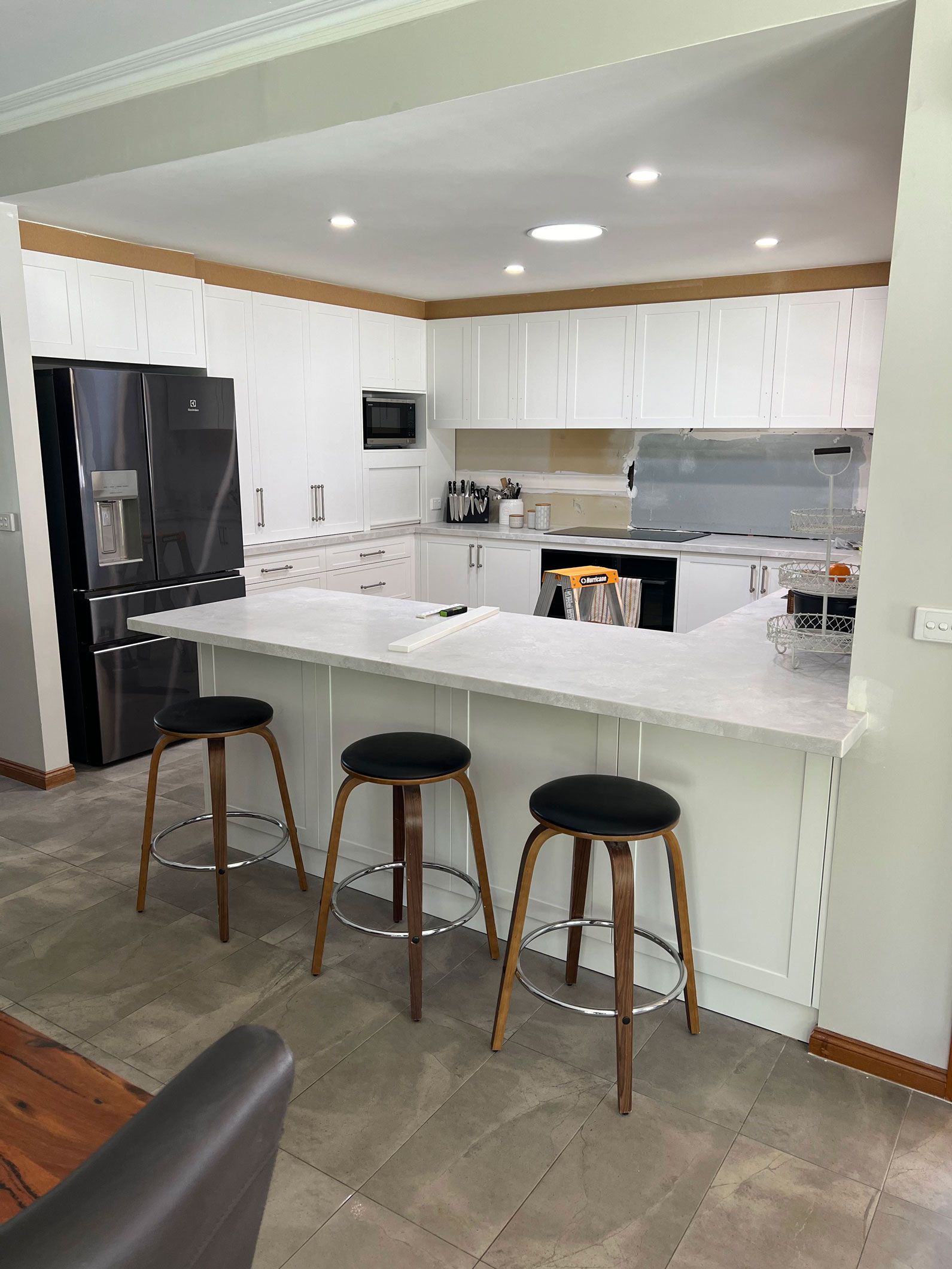Modern kitchen with white cabinets, countertop, and stools. Black refrigerator, microwave, and recessed lighting.