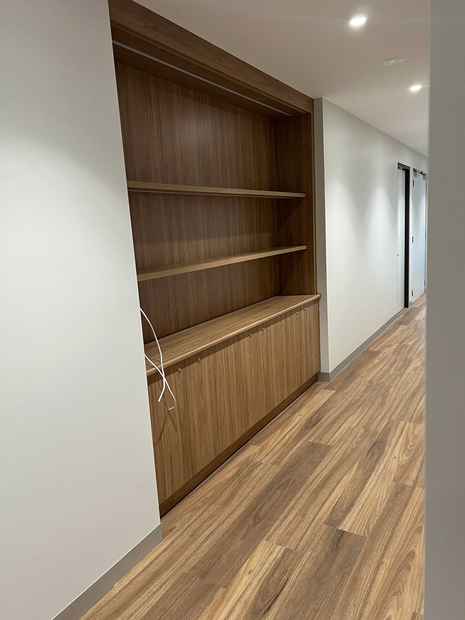Wooden built-in shelves and cabinets along a hallway with wood-look flooring and white walls.