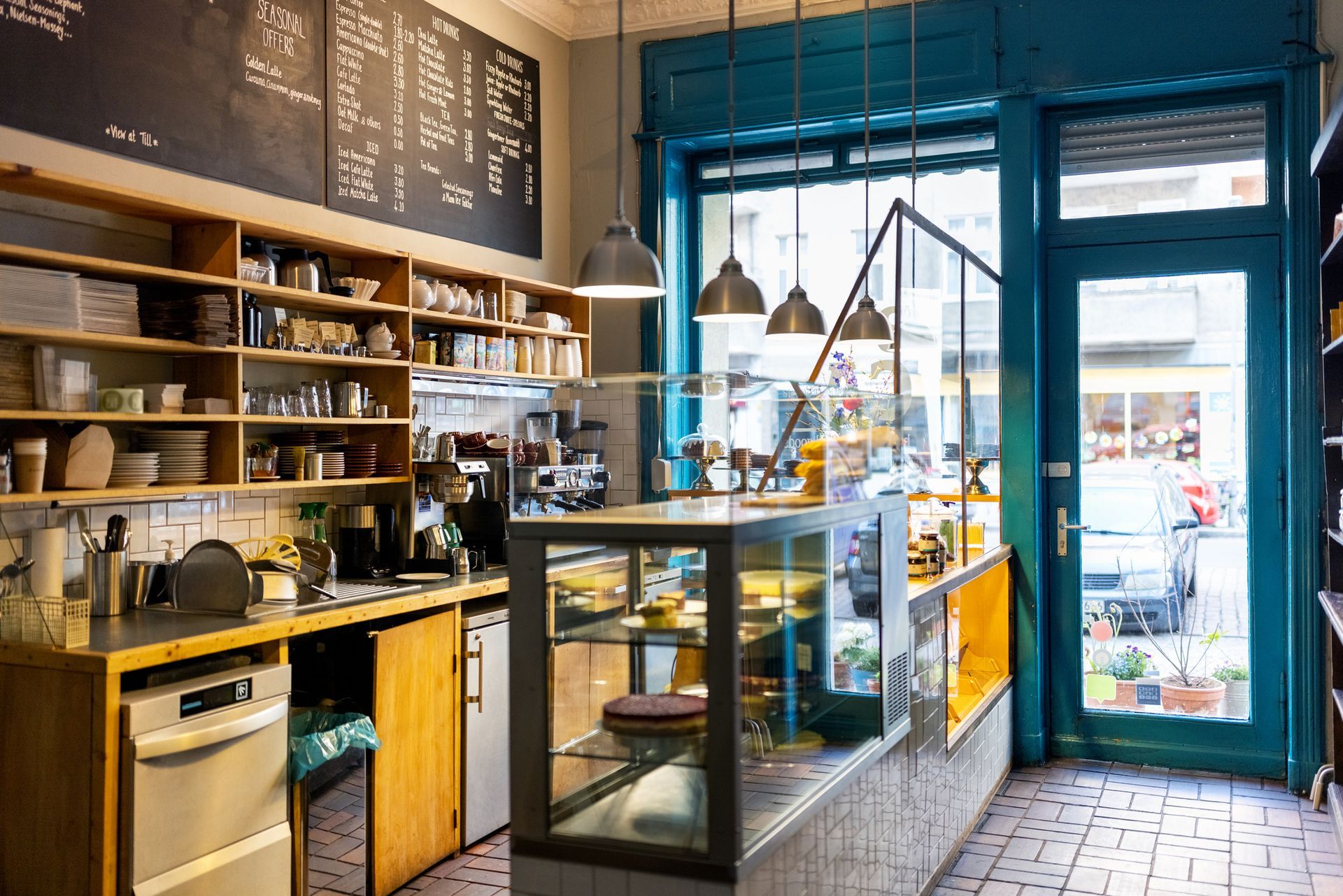 Interior of a cafe with a counter, baked goods, shelves, and a chalkboard menu. Blue and yellow accents.