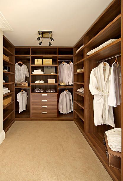 Walk-in closet with dark wood shelving, drawers, and hanging rods. Cream-colored clothes and bathrobe visible. Beige carpet.
