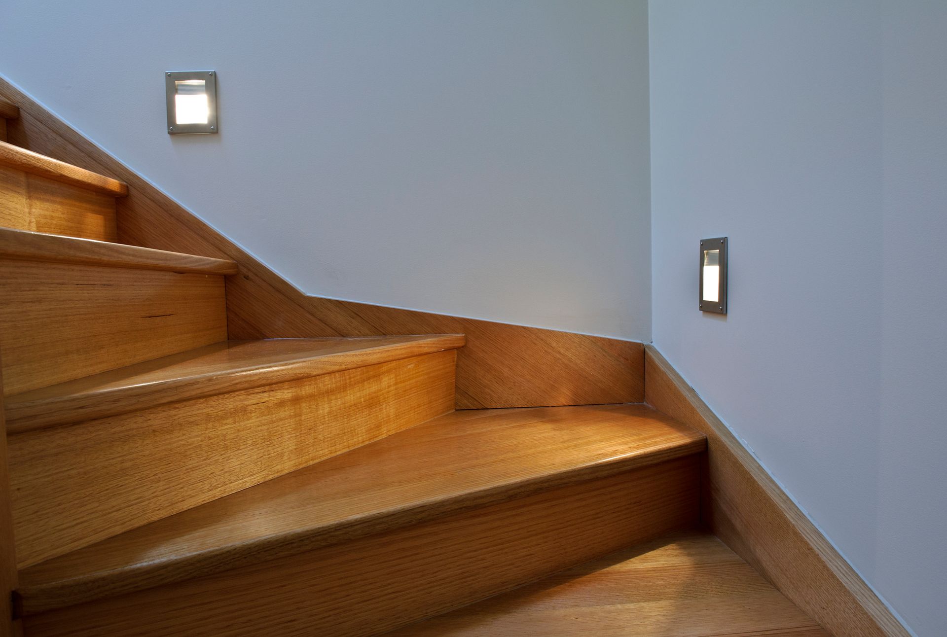 Wooden staircase with two wall-mounted lights illuminating the steps and white walls.