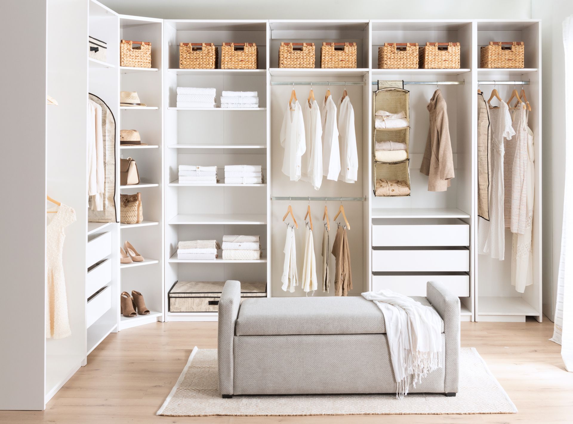 Walk-in closet with white shelving, hanging clothes, woven baskets, and a bench.