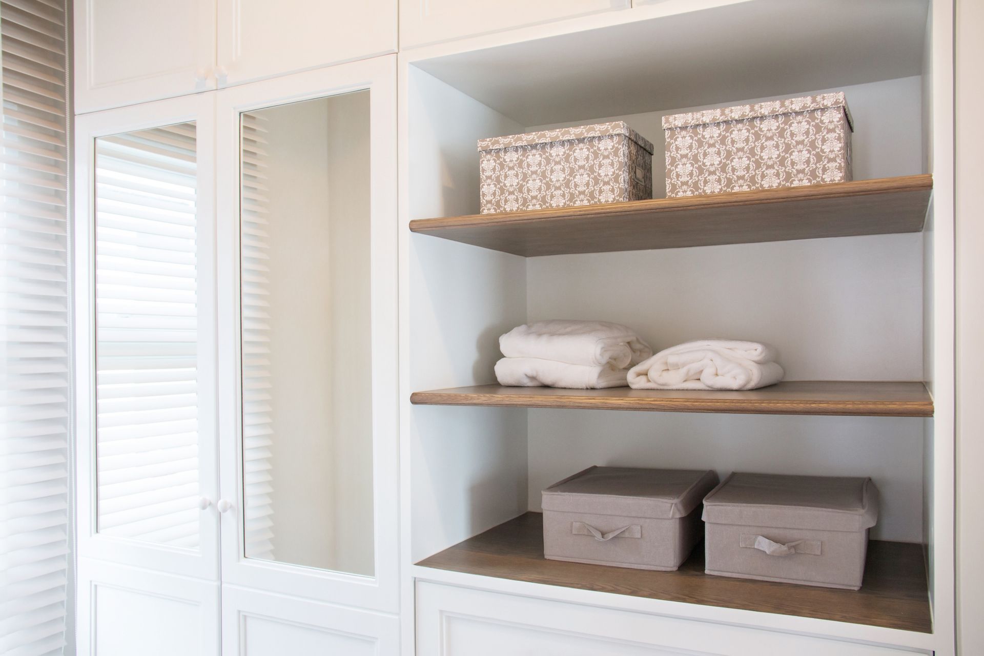 White built-in shelves with wooden shelves. Two decorative boxes, folded linens, and two gray storage boxes are visible.