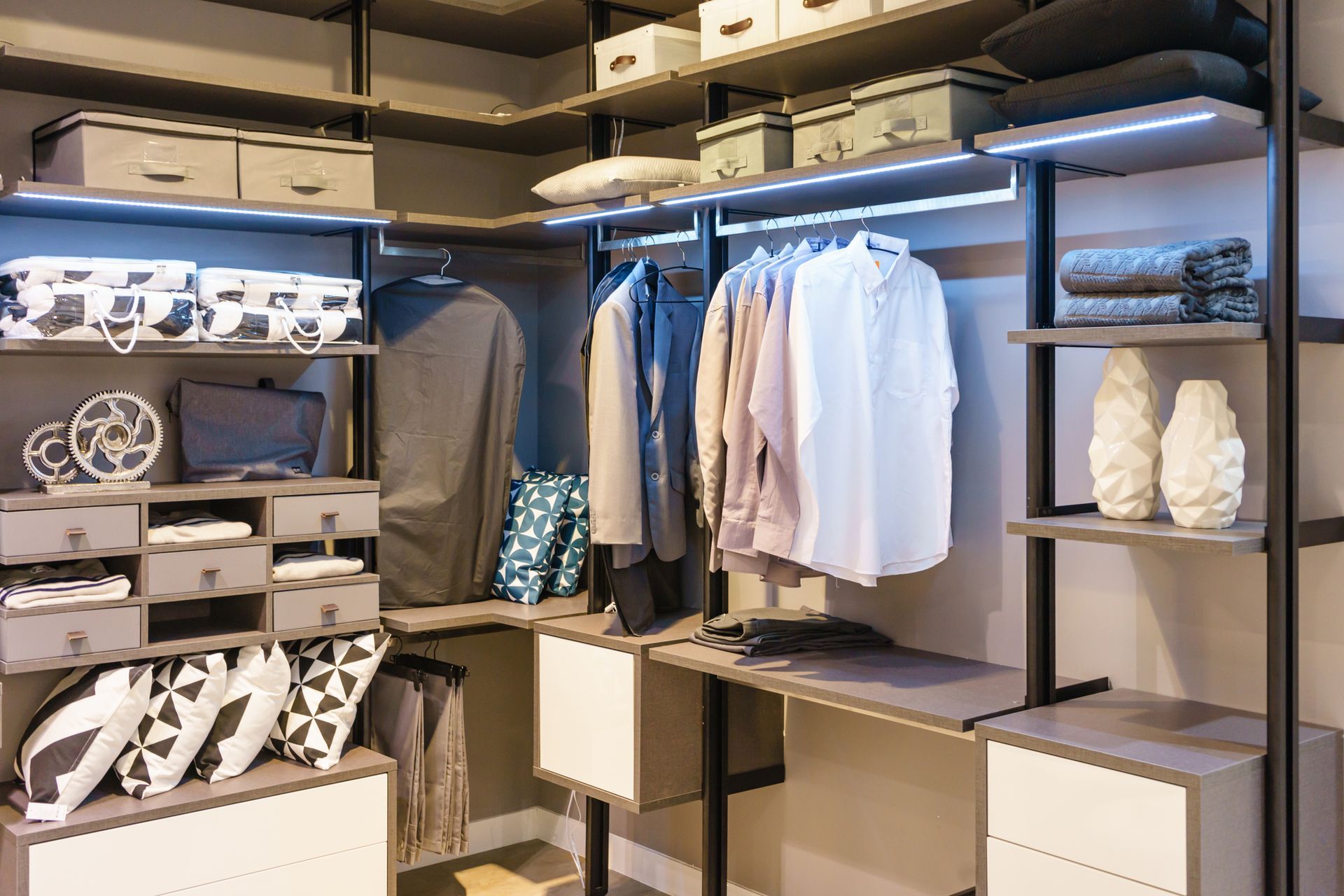 Walk-in closet with gray and white shelving, clothes, and storage boxes. Lit by LED lights.