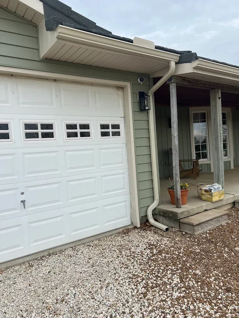 A white garage door sits next to a covered porch with a stone step and a gravel driveway on a home with green siding.