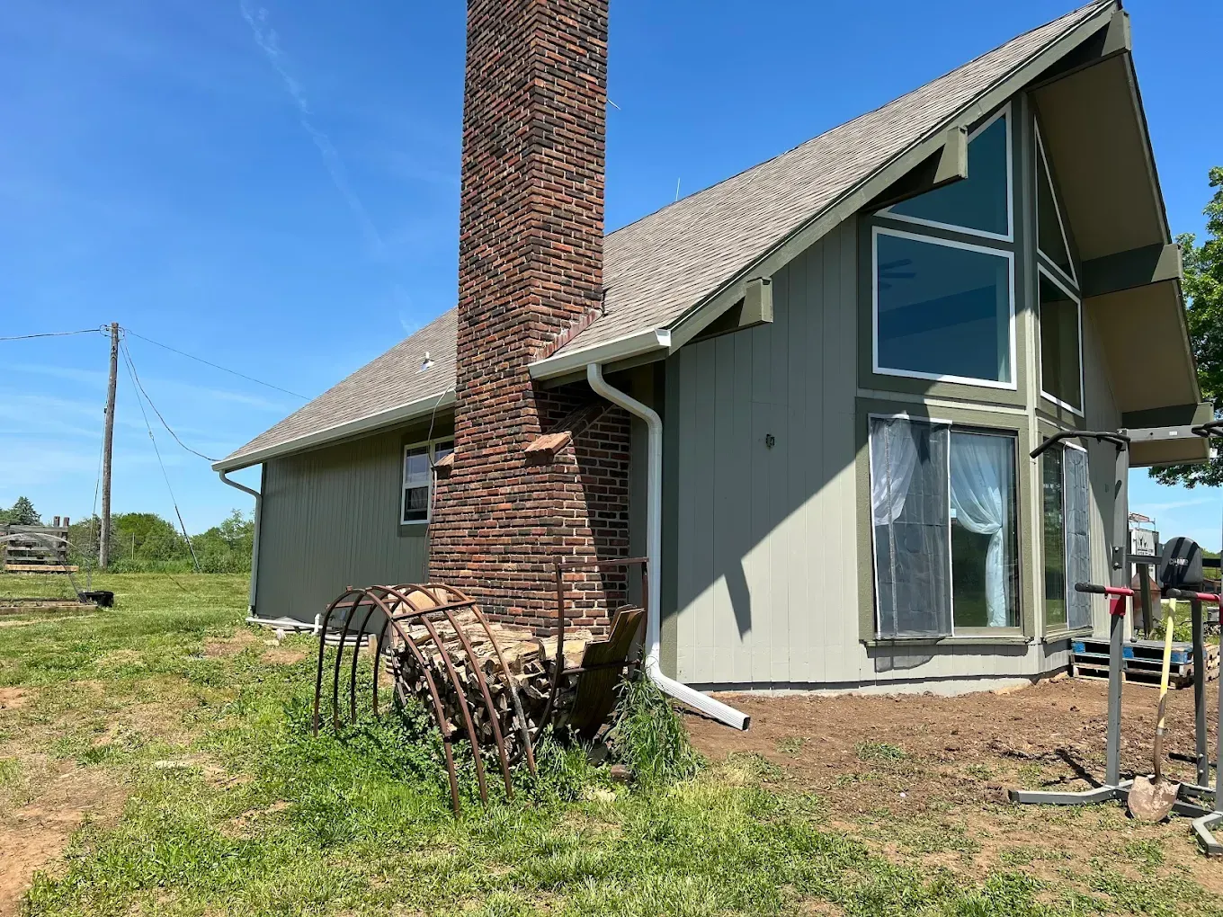 A gray, A-frame house features a prominent brick chimney and a large, rusted metal farm implement resting at its base.