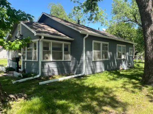 A single-story, grey-sided residential house with a shingled roof, white trim, and white downspouts on a grassy lot.