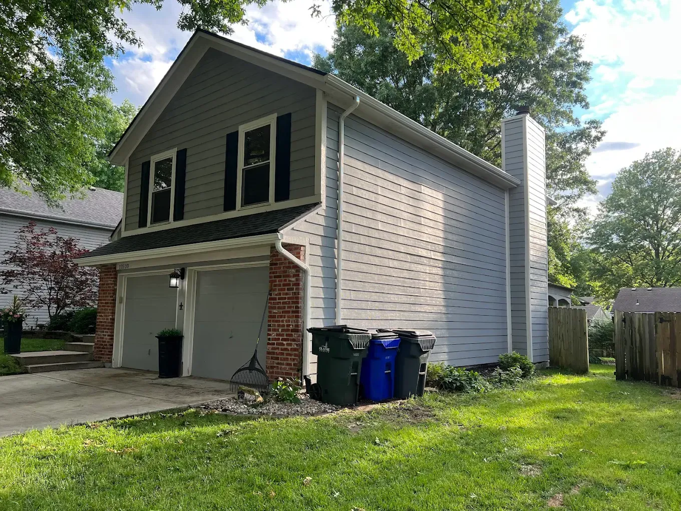 A two-story garage with grey siding, brick trim, a chimney, and three trash bins, located on a grassy residential lot.
