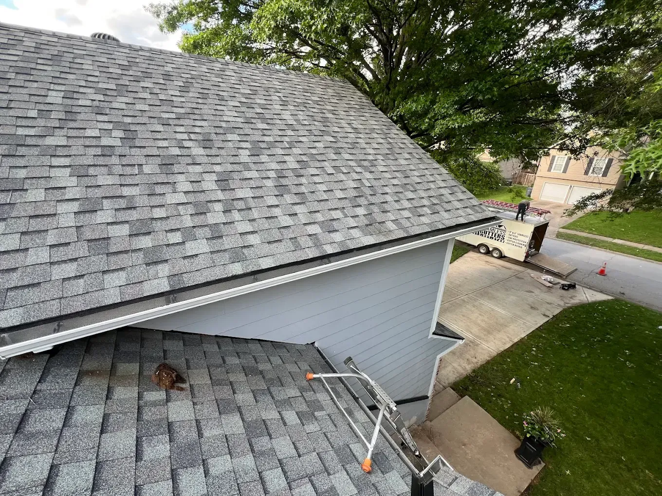 A high-angle view of a gray asphalt shingle roof with gutters, a ladder, and a nearby house and driveway.