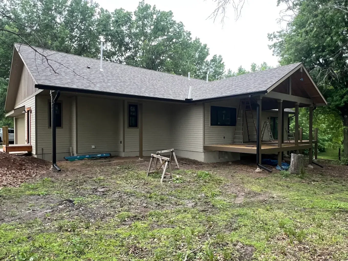 A single-story tan house with a dark roof and a covered wooden deck, set against a wooded background.