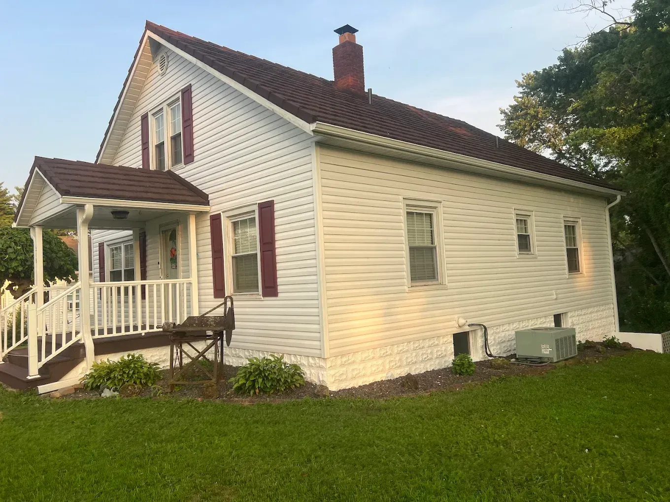 A white, one-story house with a brown shingled roof, red shutters, and a front porch, set on a grassy lawn.