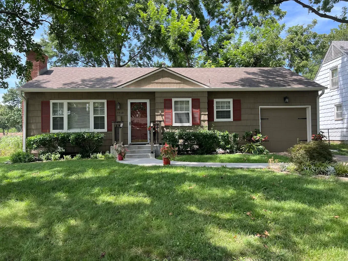 A single-story brown house with red shutters, a front door under a small gable, and a single-car garage under a blue sky.