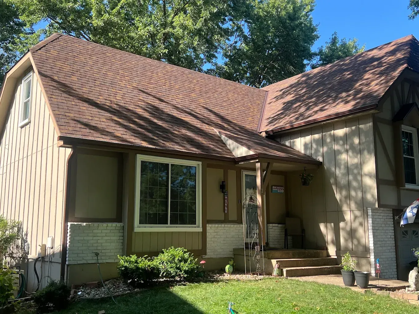 A beige Tudor-style home with brown shingles, stone accents, and a small front porch, set against a green lawn and trees.