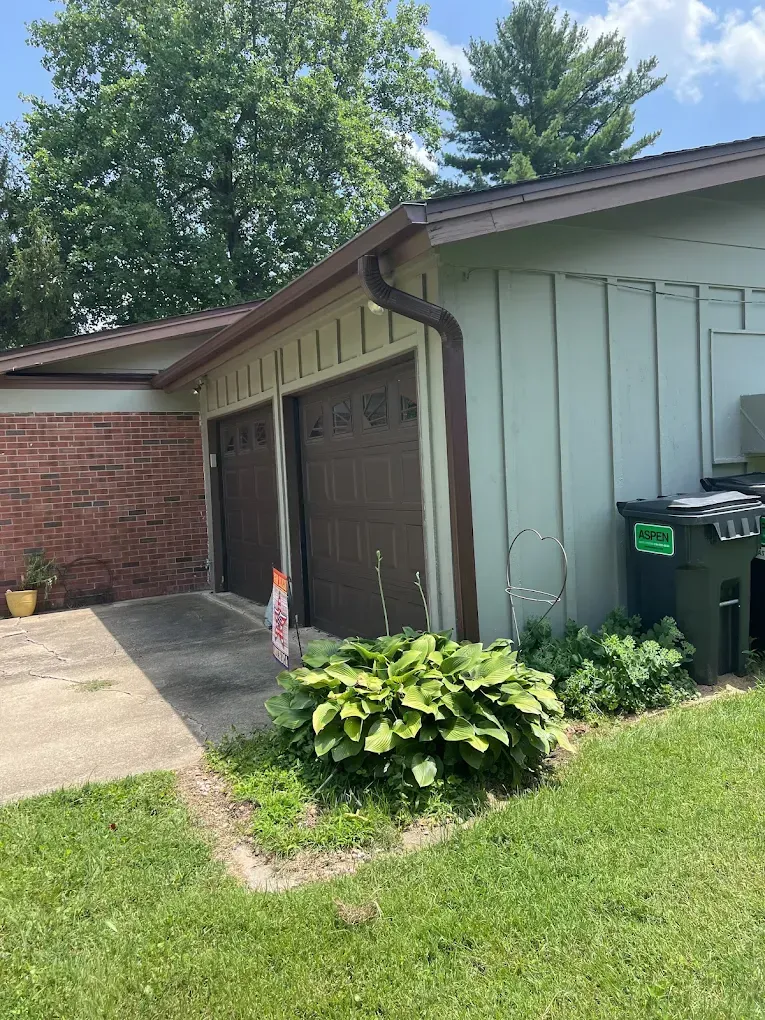 A view of a garage with brown doors, a brick wall, green siding, and a large bush in the front yard.