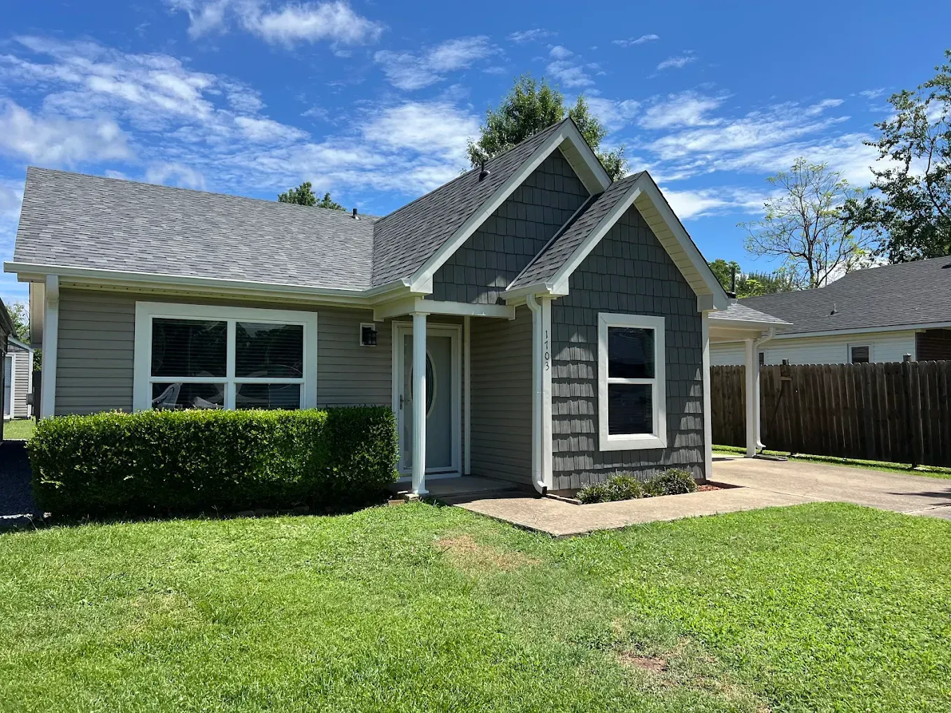 A single-story gray house with a gable roof, dark shingle siding on the entryway, and a lush green lawn.