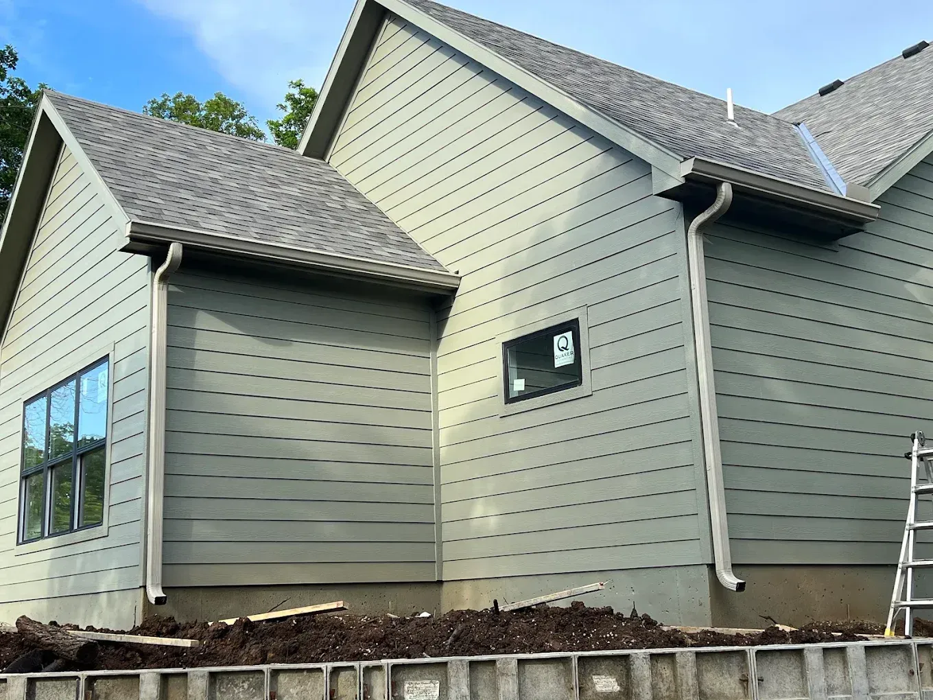 Sage green house with horizontal lap siding and two downspouts against a backdrop of trees and a dirt foundation.