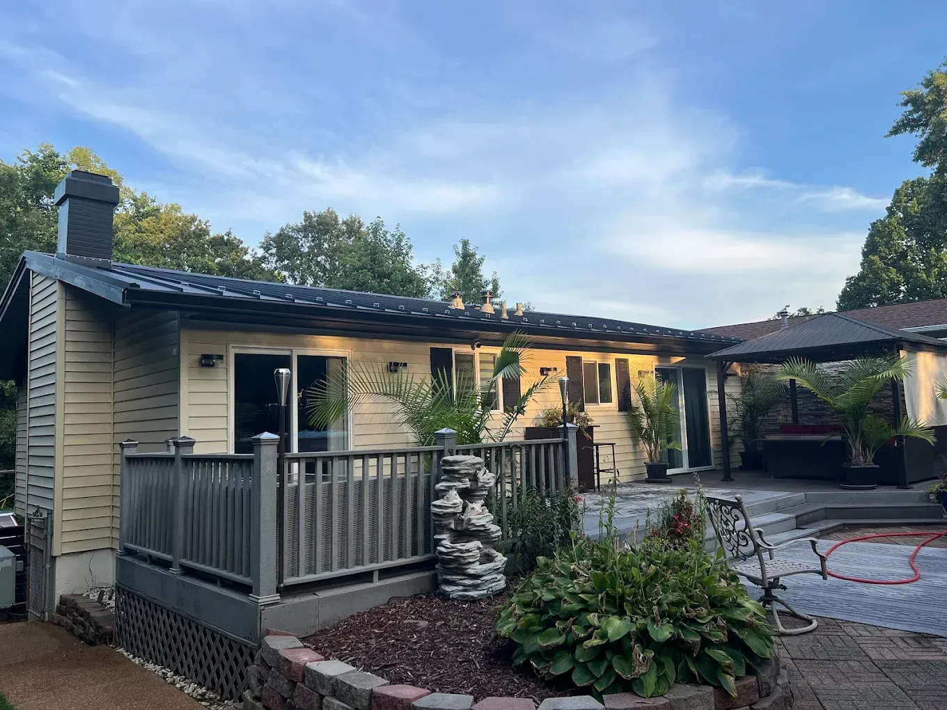 A beige house with a grey deck, a stone water feature, and a patio area with a gazebo under a blue sky.